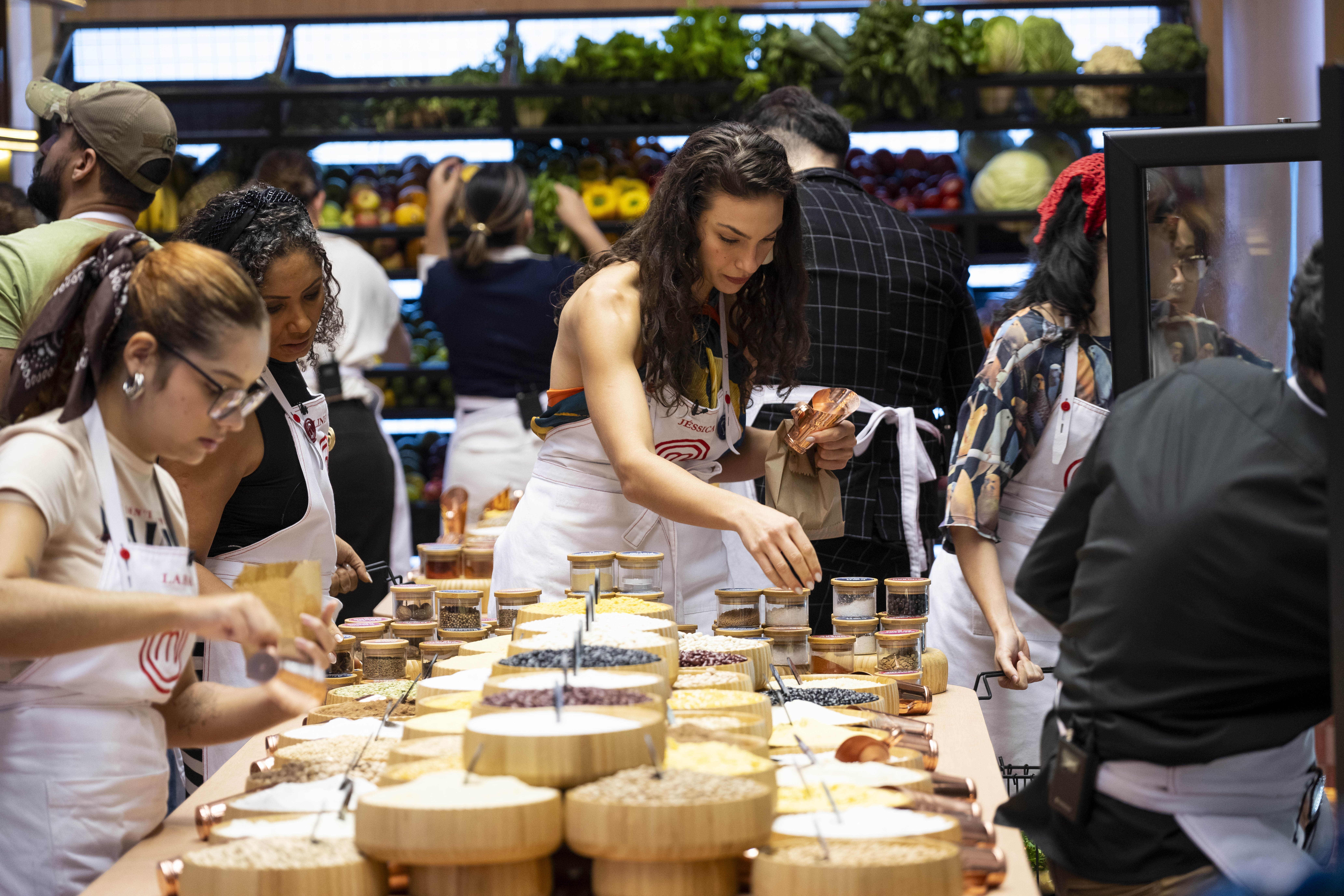 Mulheres com aventais brancos preparam ingredientes em uma bancada cheia de potes e tigelas com gr&atilde;os e temperos. Ao fundo, outras pessoas e prateleiras com frutas e vegetais frescos em um mercado ou supermercado