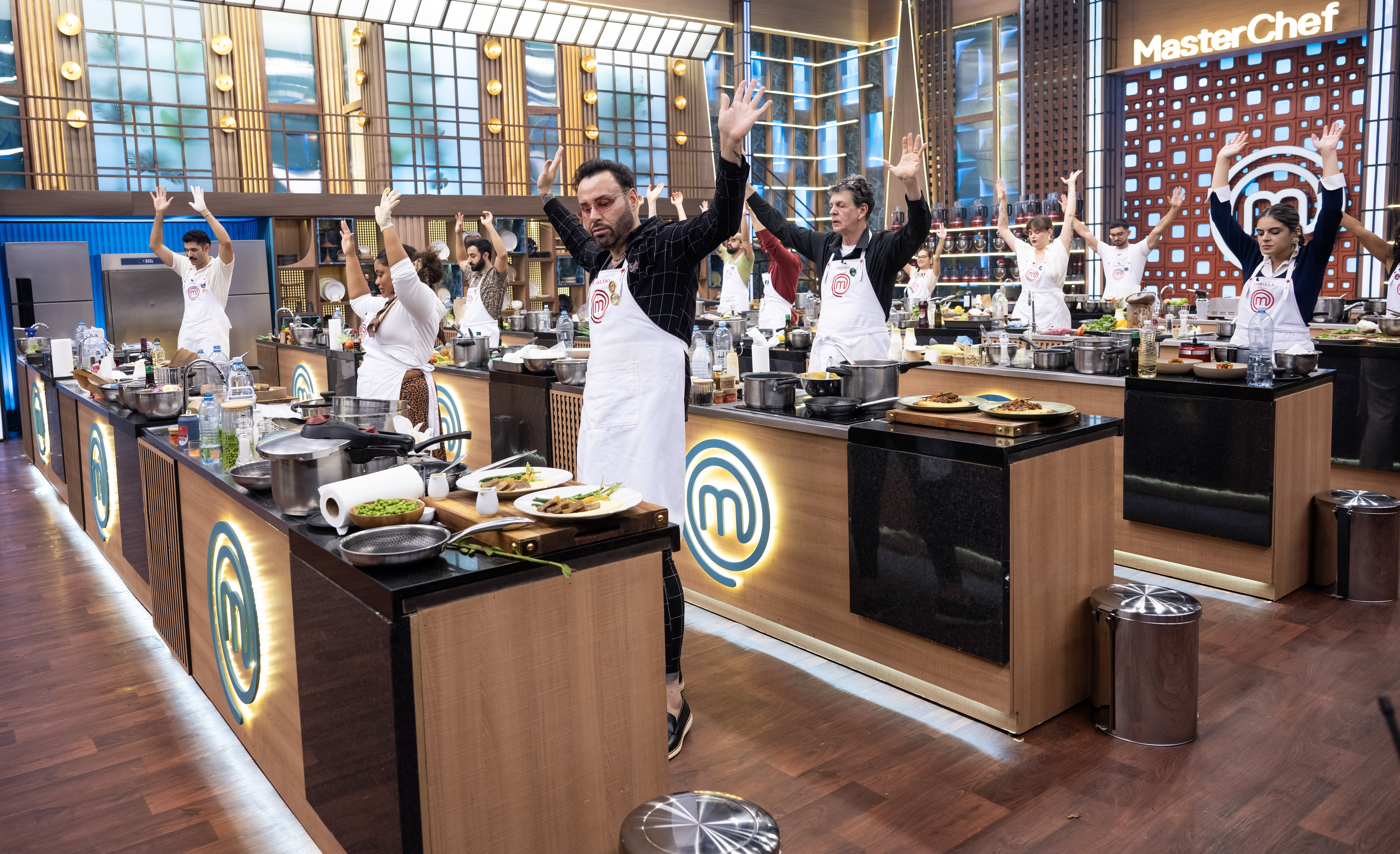 Competidores do MasterChef, usando aventais brancos, levantam os bra&ccedil;os em comemora&ccedil;&atilde;o ou instru&ccedil;&atilde;o, em uma cozinha profissional com bancadas de madeira e metal, panelas e pratos prontos. Ao fundo, o logo do programa.