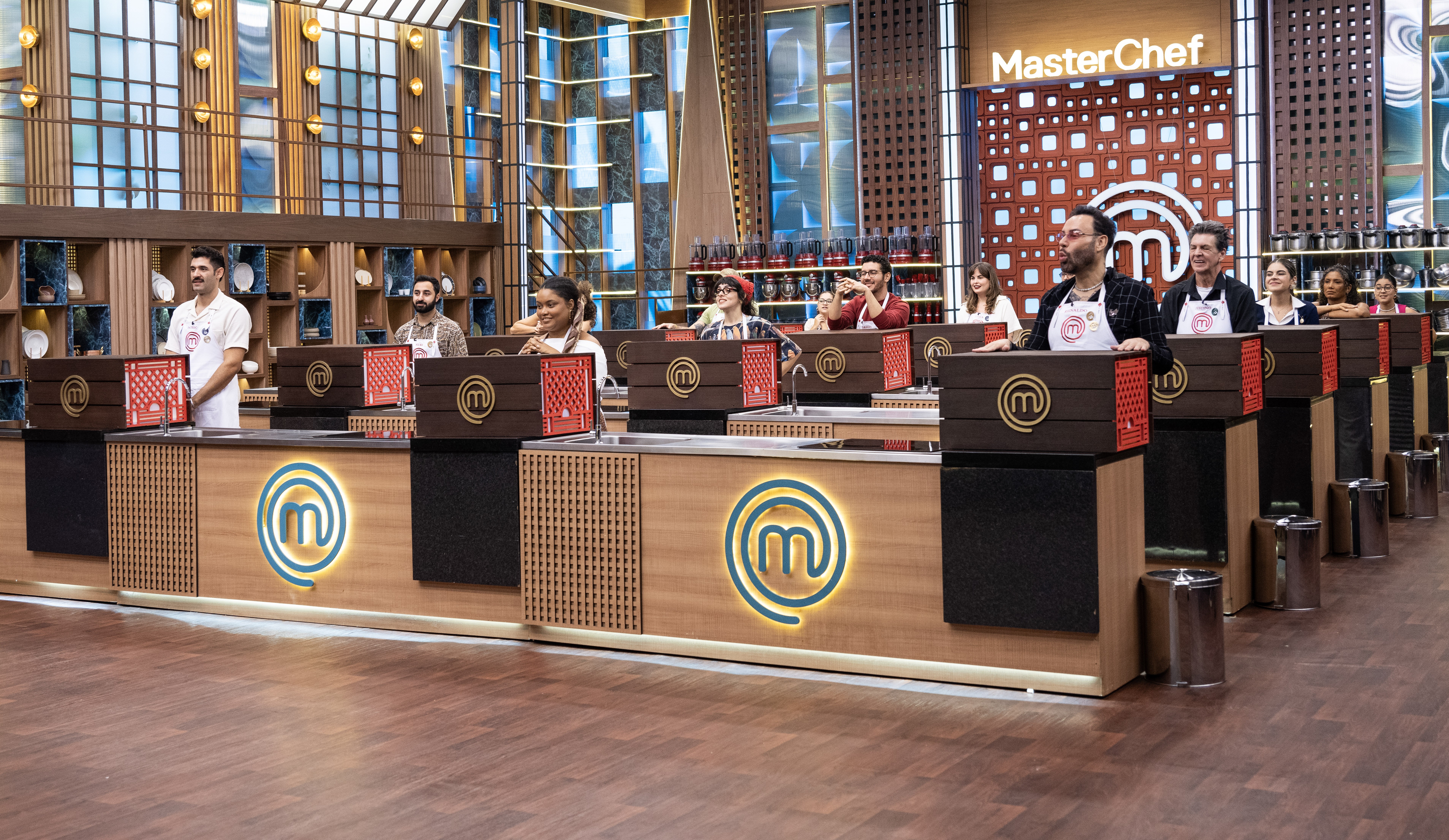 Competidores do MasterChef Brasil, homens e mulheres de diversas etnias, em p&eacute; atr&aacute;s de bancadas de madeira com o logo M em neon azul, no est&uacute;dio do programa