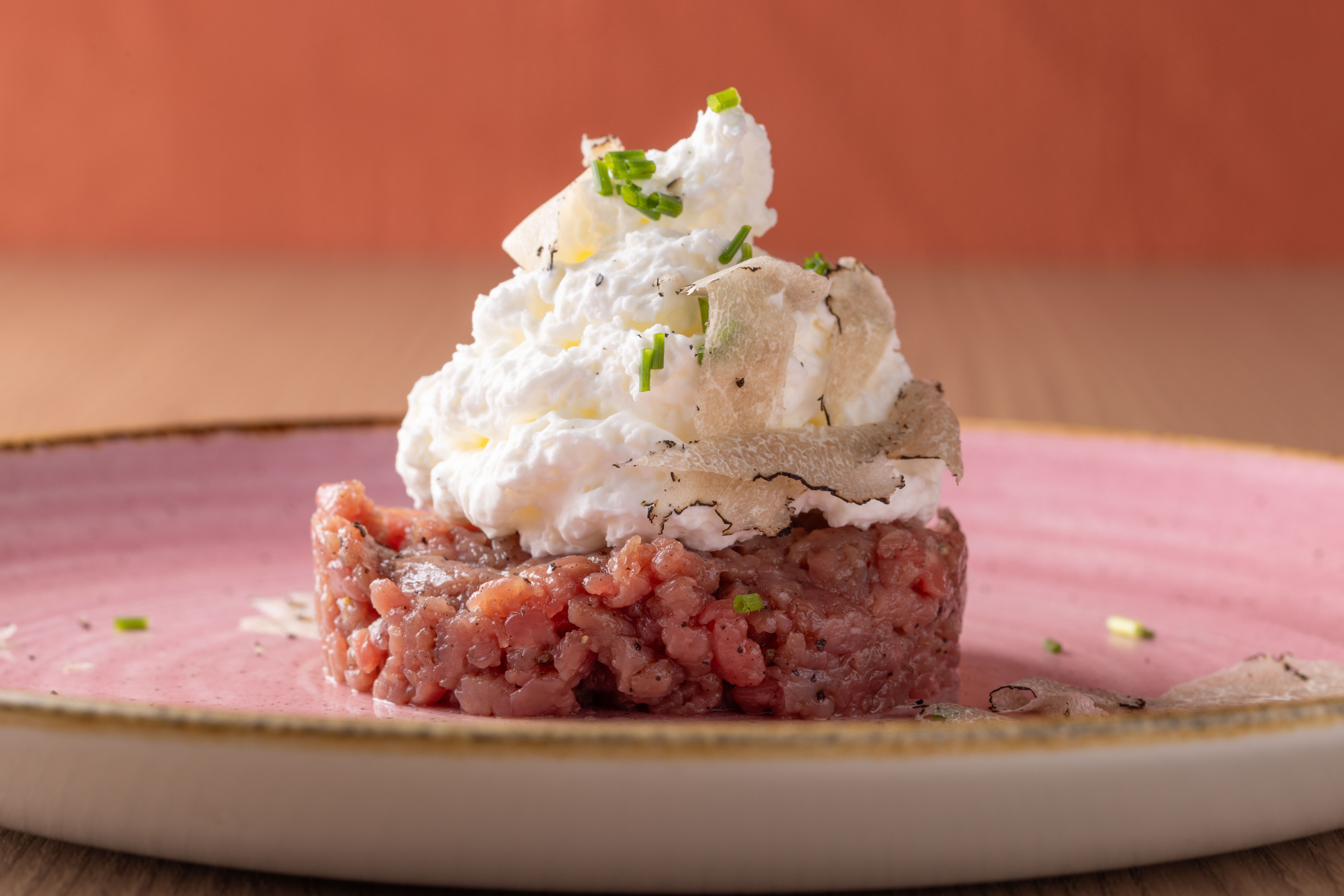 Tartare de carne vermelha mo&iacute;da, coberto com creme branco, fatias finas de trufa e cebolinha picada, servido em um prato rosa com borda dourada