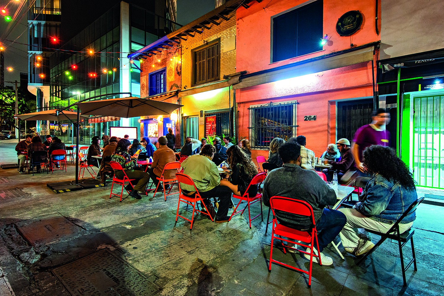 Pessoas sentadas em mesas com cadeiras vermelhas na cal&ccedil;ada de um bar &agrave; noite, sob guarda-s&oacute;is e luzes coloridas penduradas. Ao fundo, pr&eacute;dios iluminados e um letreiro com o n&uacute;mero 264