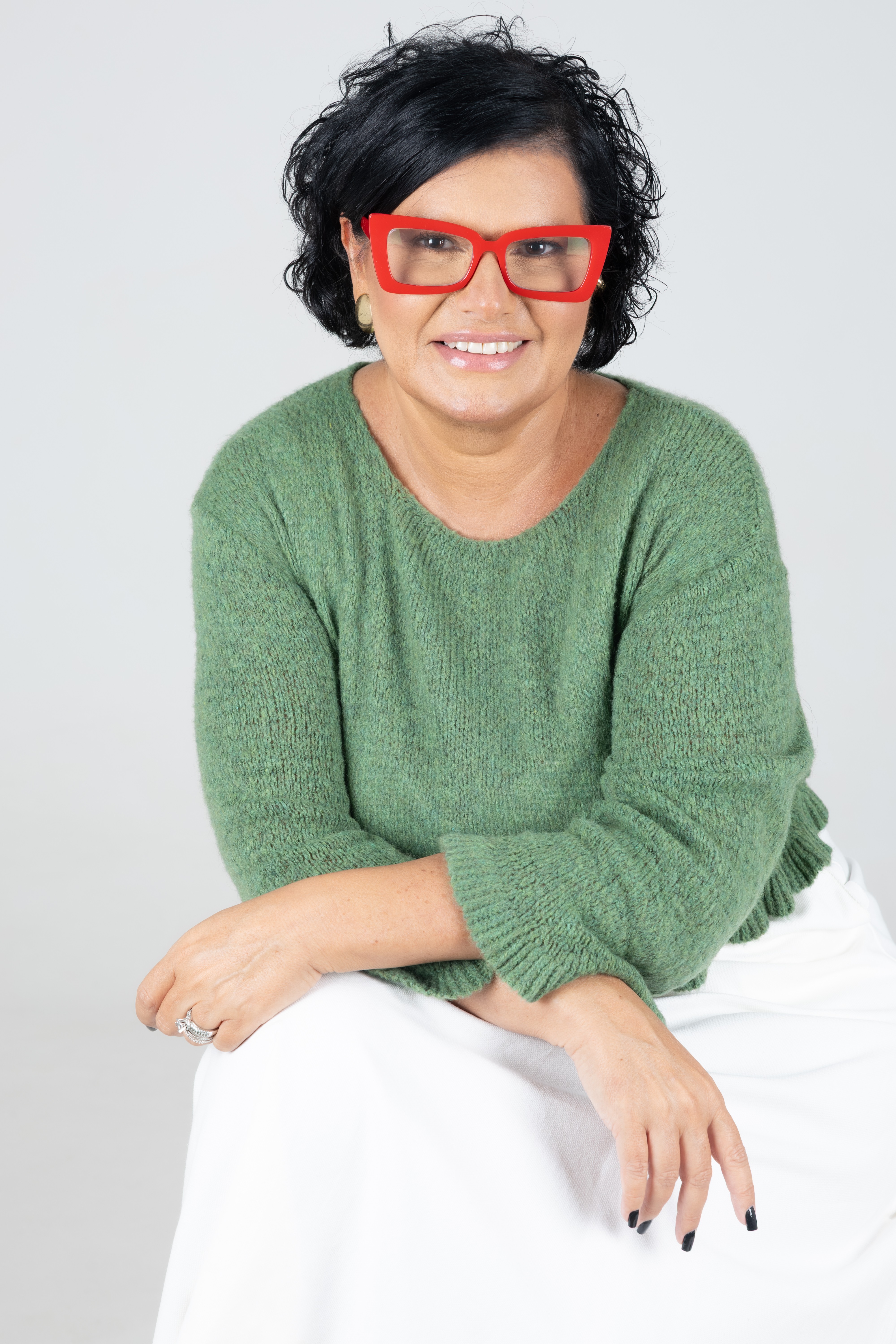 Mulher de meia-idade, cabelos pretos curtos e cacheados, sorrindo, usando &oacute;culos vermelhos grandes, su&eacute;ter verde e saia branca, sentada com as m&atilde;os cruzadas sobre as pernas