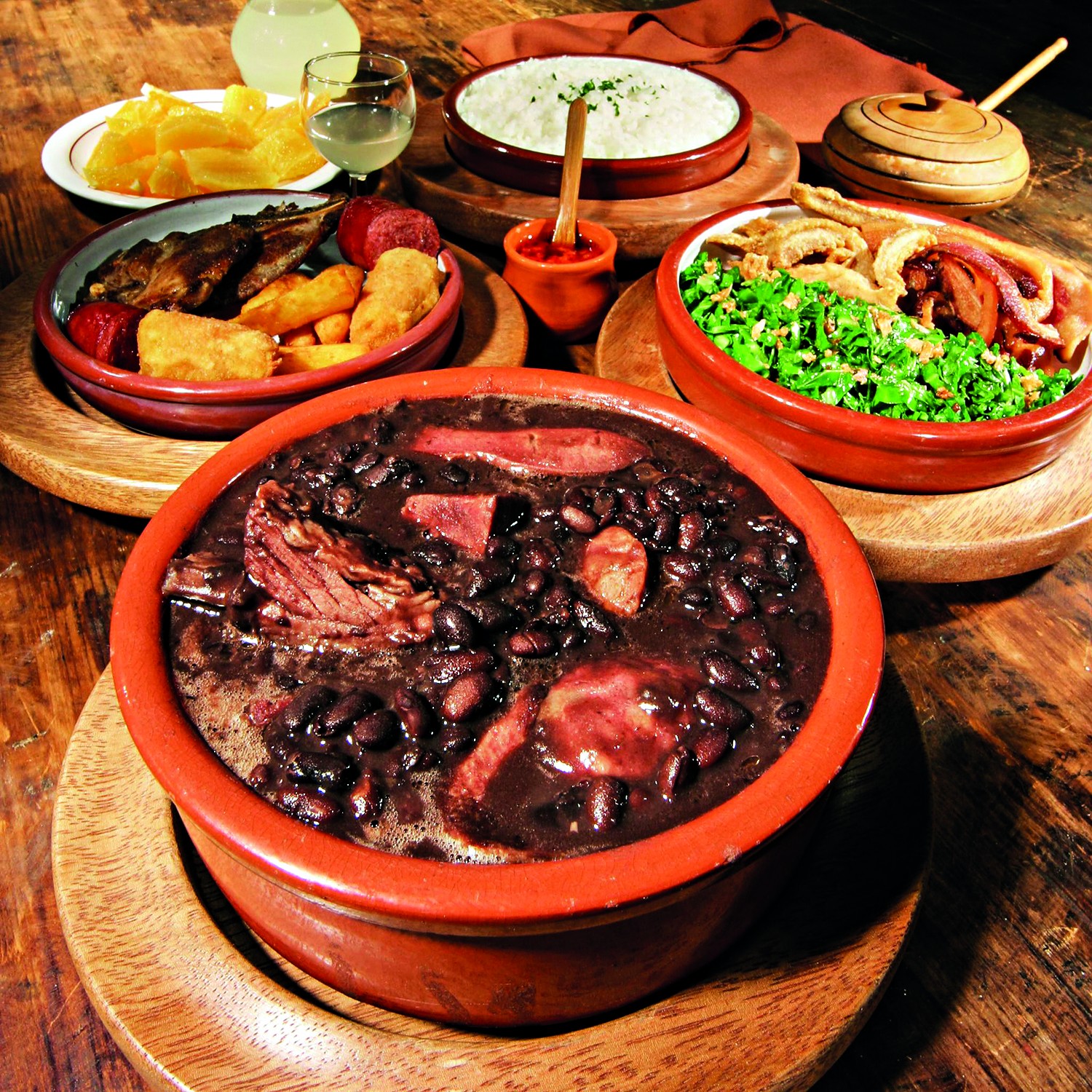 Mesa de madeira com feijoada completa em panelas de barro: feij&atilde;o preto com carnes, arroz branco, couve refogada com torresmo, farofa, lingui&ccedil;a, mandioca frita e caipirinha
