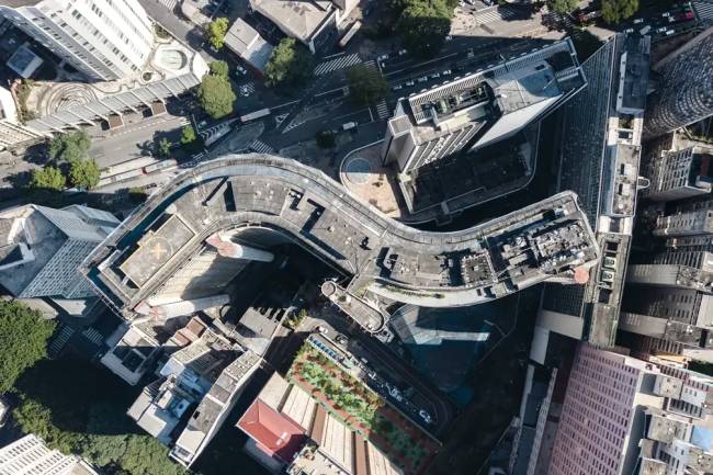 Vista a&eacute;rea de uma cidade com edif&iacute;cios e ruas. Um pr&eacute;dio curvo com telhado cinza e um heliponto no centro, cercado por outros edif&iacute;cios de diferentes alturas e formatos. Ruas com carros e &ocirc;nibus, e algumas &aacute;reas verdes com &aacute;rvores. A luz do sol cria sombras alongadas, indicando um dia claro