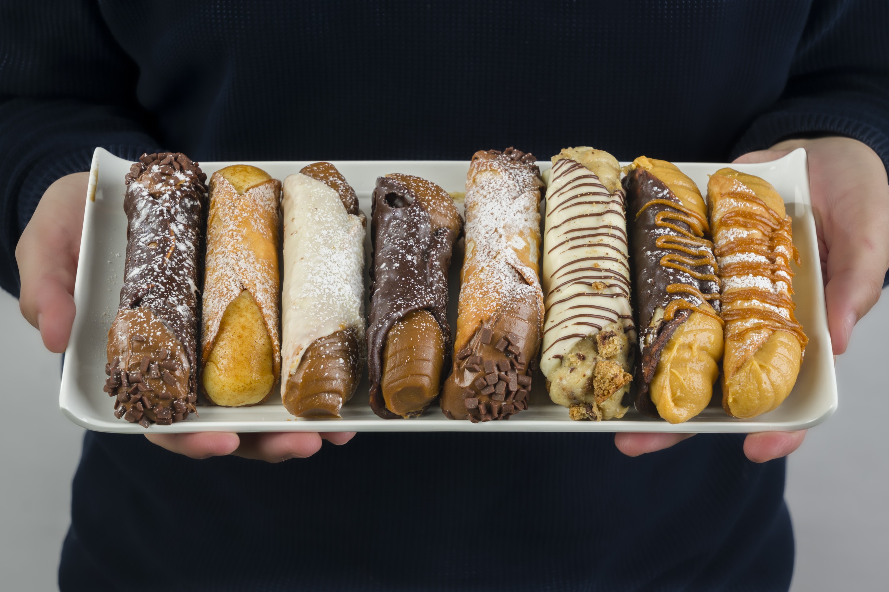 Pessoa segurando uma bandeja branca com oito cannolis de diferentes sabores, incluindo chocolate, doce de leite e cobertura de a&ccedil;&uacute;car de confeiteiro