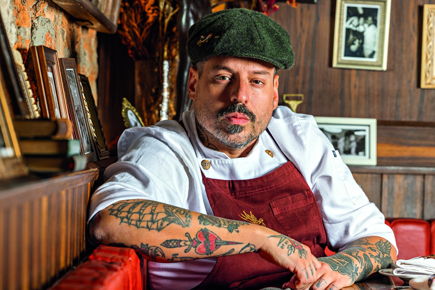 Homem de barba e bigode, usando boina verde e avental vinho sobre d&oacute;lm&atilde; branco, com bra&ccedil;os tatuados apoiados em um sof&aacute; vermelho, olhando para frente. Ao fundo, parede de madeira com quadros e objetos decorativos