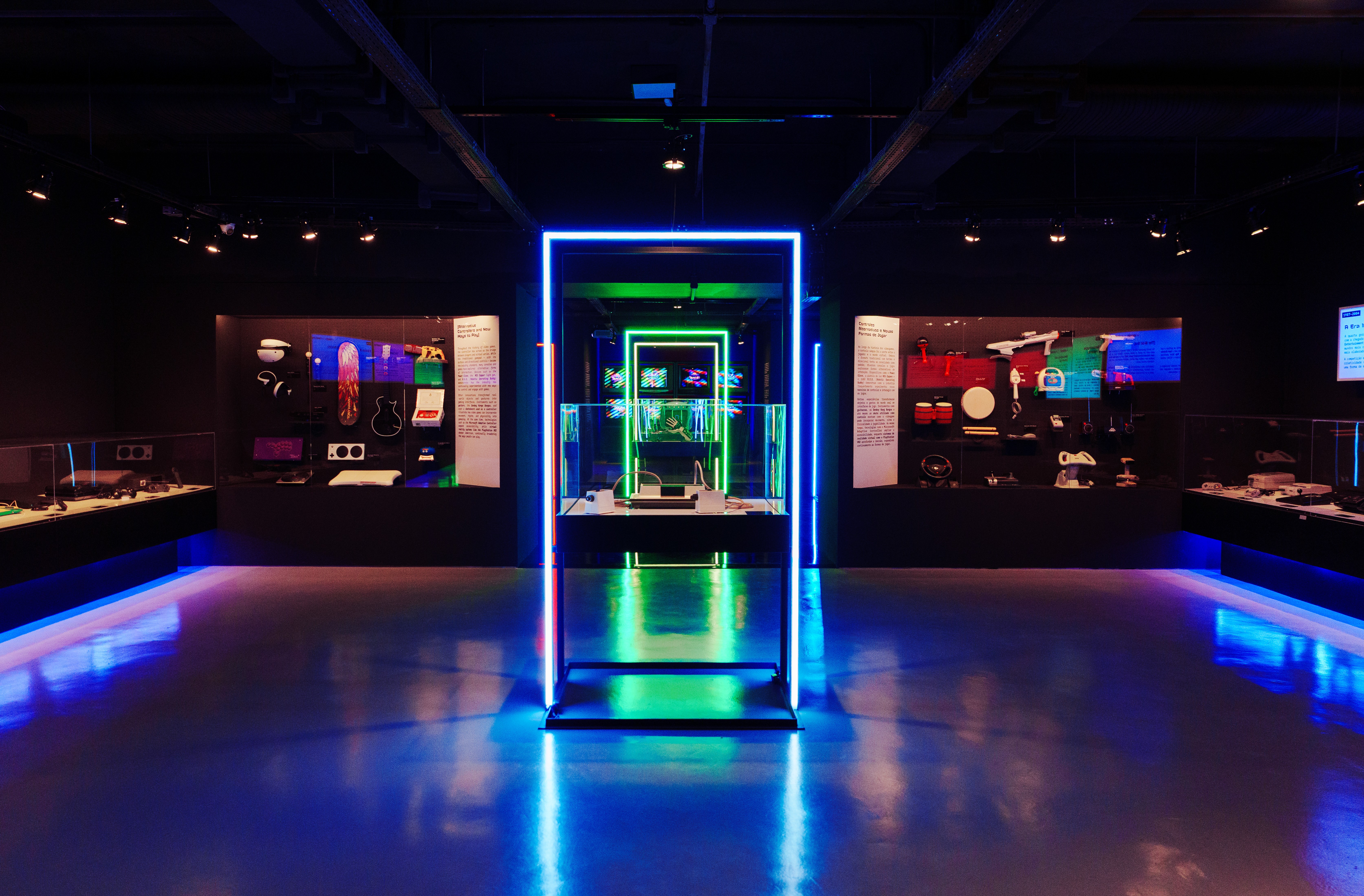 Exposi&ccedil;&atilde;o futurista com ilumina&ccedil;&atilde;o neon azul e verde em um ambiente escuro. No centro, uma vitrine com um objeto tecnol&oacute;gico &eacute; emoldurada por um ret&acirc;ngulo de luz neon azul, e outro ret&acirc;ngulo verde menor ao fundo. Nas laterais, pain&eacute;is expositivos com objetos e textos informativos. O ch&atilde;o reflete as luzes, criando um efeito vibrante e moderno