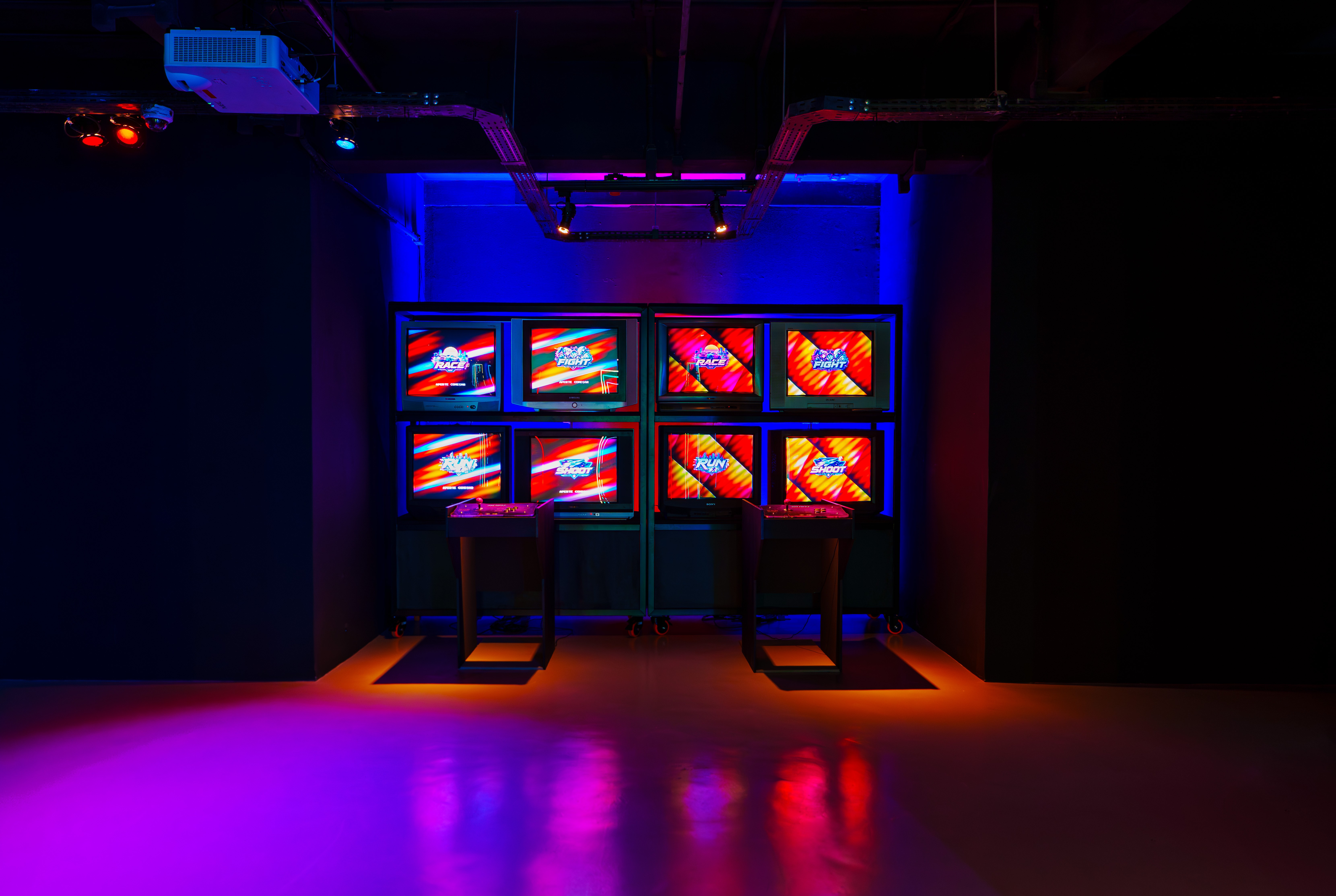 Sala escura com oito monitores de tubo exibindo a mesma imagem colorida e abstrata, dispostos em duas colunas de quatro. Dois pedestais com controles de videogame est&atilde;o &agrave; frente, um em cada lado. O ambiente &eacute; iluminado por luzes neon azuis e roxas, refletindo no ch&atilde;o.