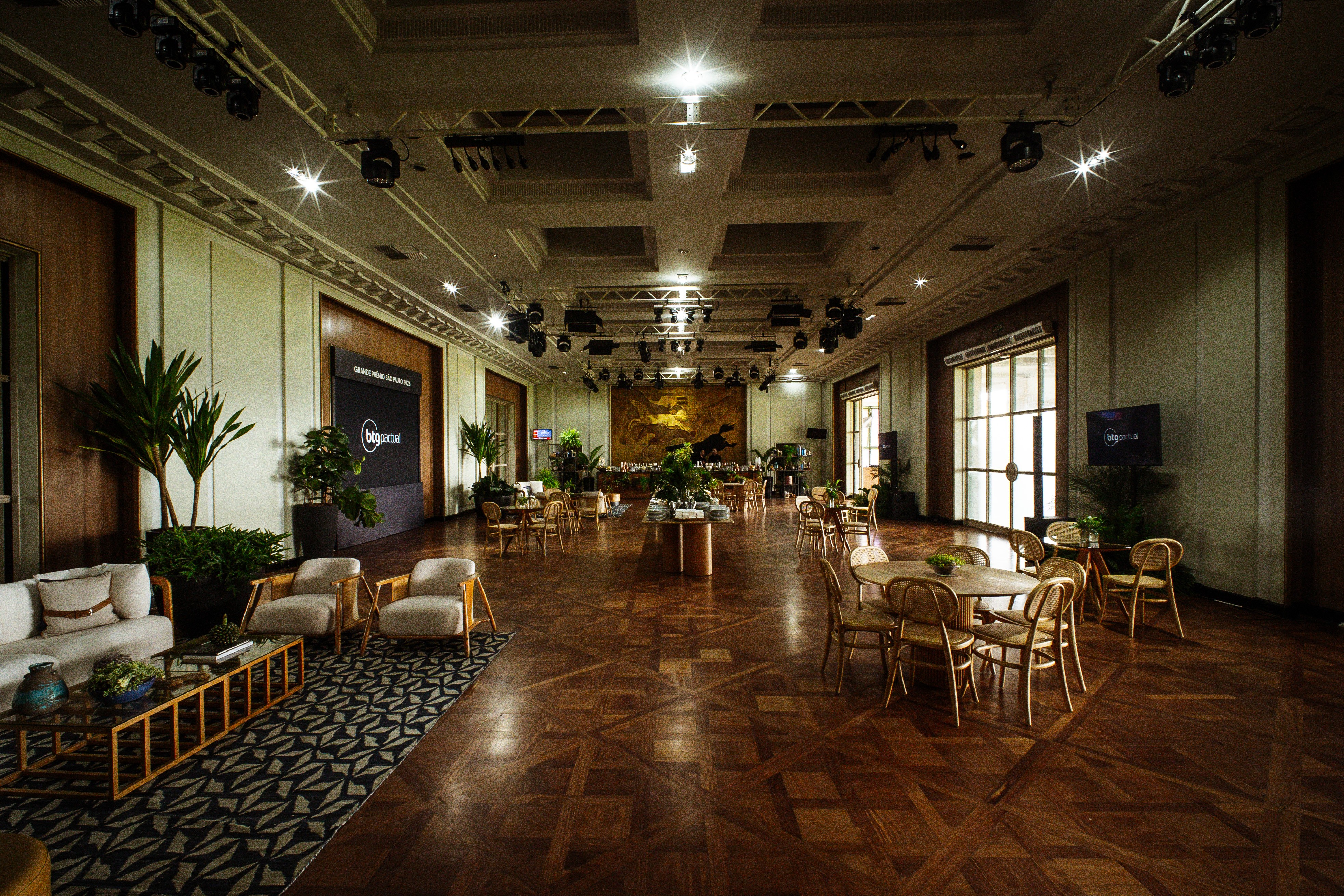 Sal&atilde;o de eventos amplo com piso de madeira, mesas redondas e cadeiras de vime, sof&aacute;s claros e poltronas, plantas verdes, ilumina&ccedil;&atilde;o focada no teto e telas pretas exibindo o logo bigportal
