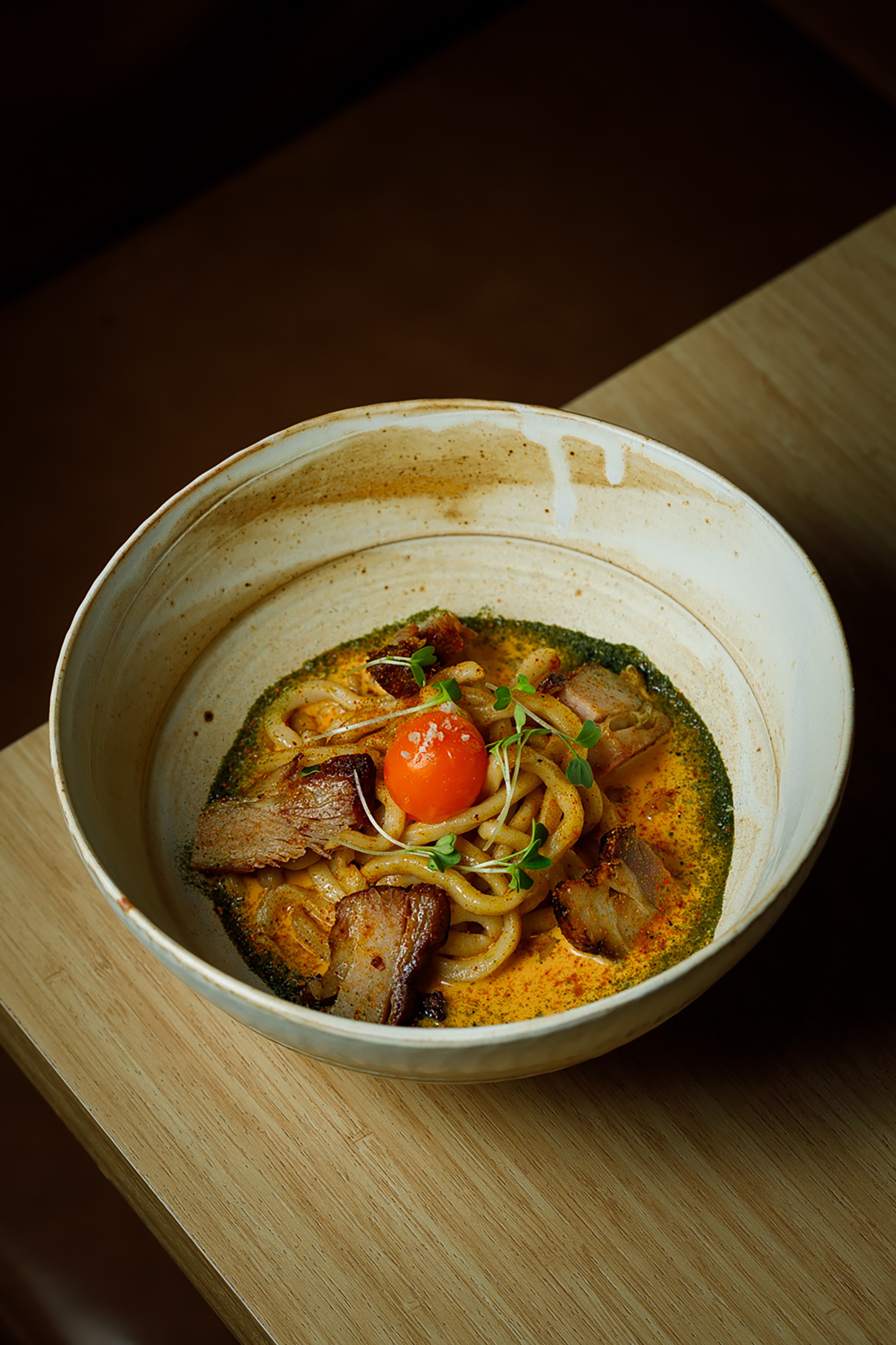Prato de massa udon com fatias de carne, molho cremoso laranja e verde, um tomate cereja e brotos frescos, servido em uma tigela r&uacute;stica de cer&acirc;mica sobre uma mesa de madeira