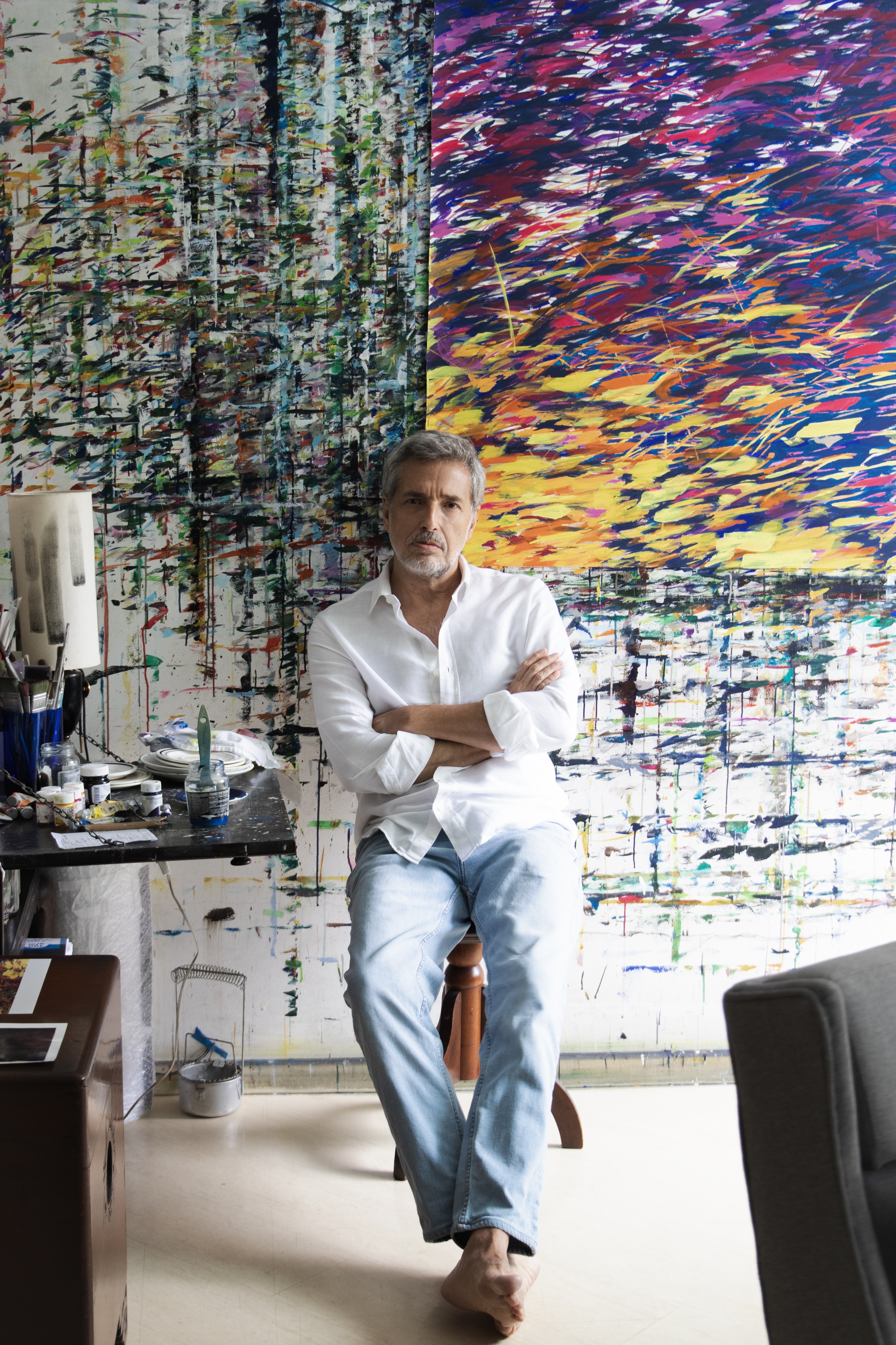 Homem de cabelos grisalhos e barba, camisa branca e jeans, sentado descal&ccedil;o em um ateli&ecirc; com quadros abstratos coloridos ao fundo e uma mesa com materiais de pintura