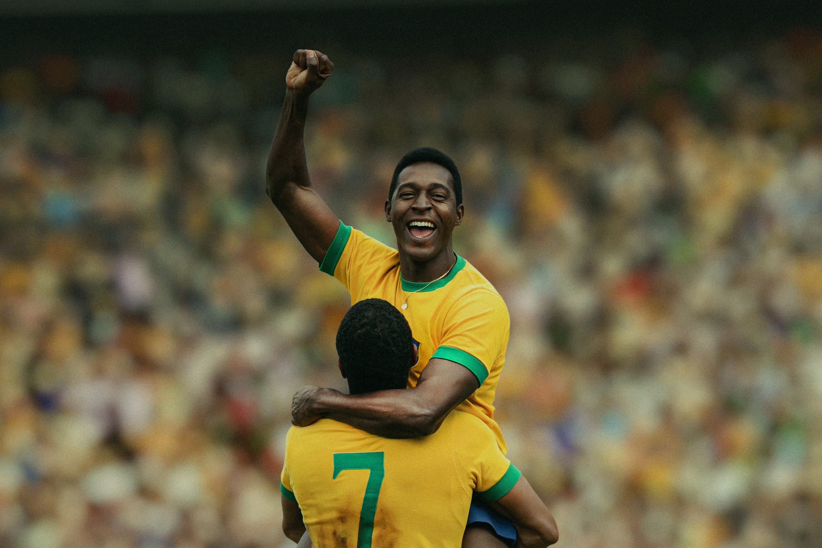 Jogador de futebol negro, sorrindo e comemorando com o punho erguido, sendo carregado por outro jogador em campo, ambos vestindo camisas amarelas com detalhes verdes, em um estádio lotado