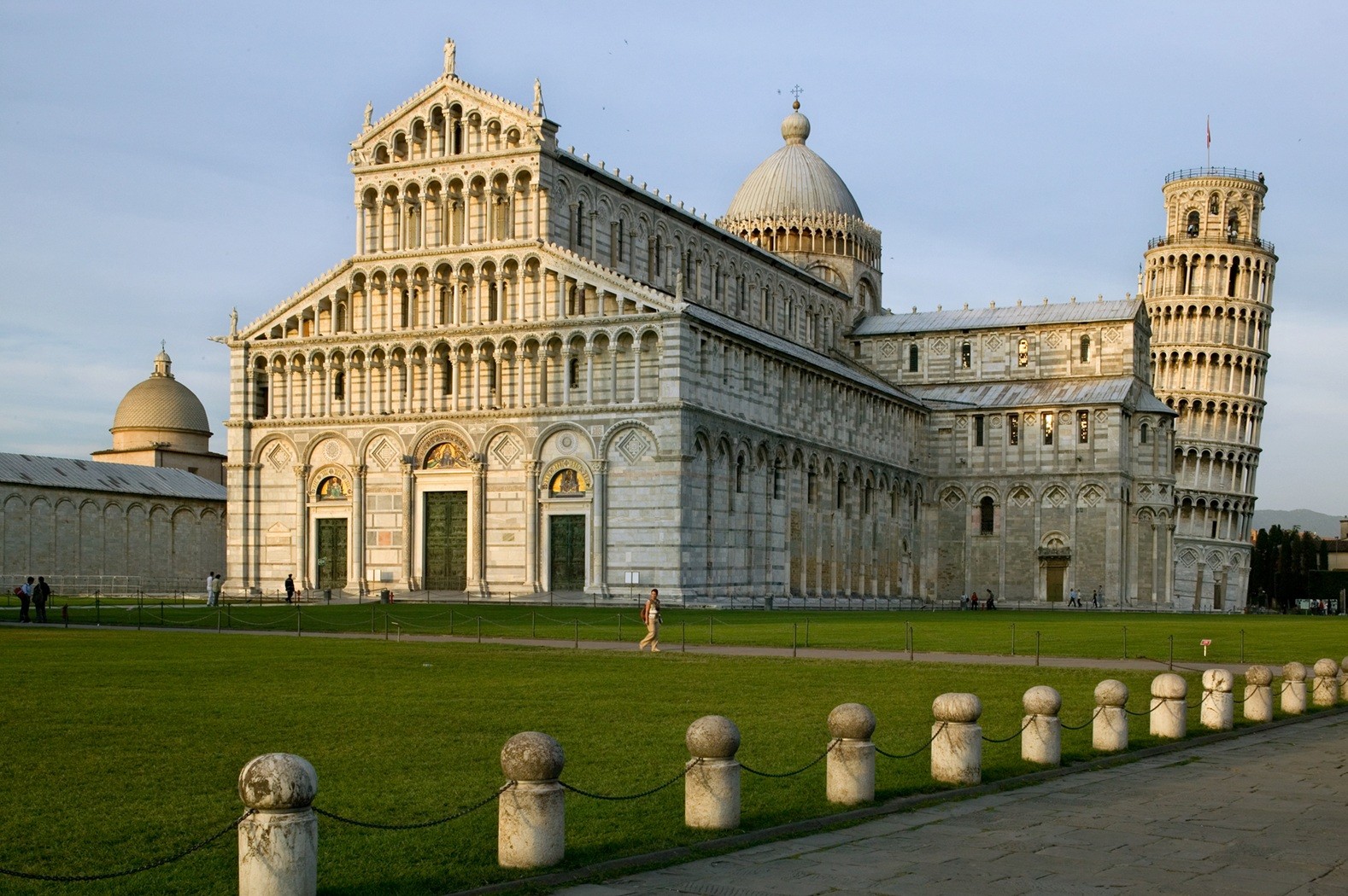 O Duomo de Pisa, uma catedral branca ornamentada, e a Torre Inclinada de Pisa, em estilo rom&acirc;nico, sob um c&eacute;u claro. Um gramado verde e um caminho de pedra com pilares baixos e correntes em primeiro plano.
