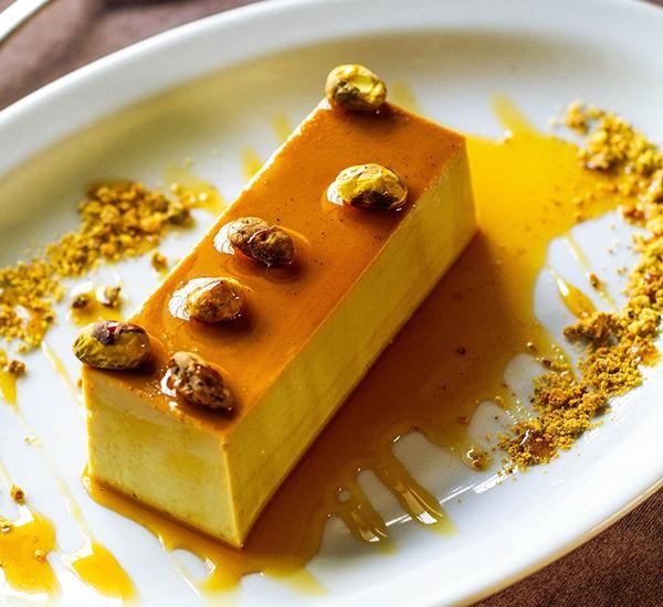 Fatia retangular de pudim amarelo claro com cobertura de caramelo brilhante, decorada com pistaches inteiros e granulado dourado em um prato branco