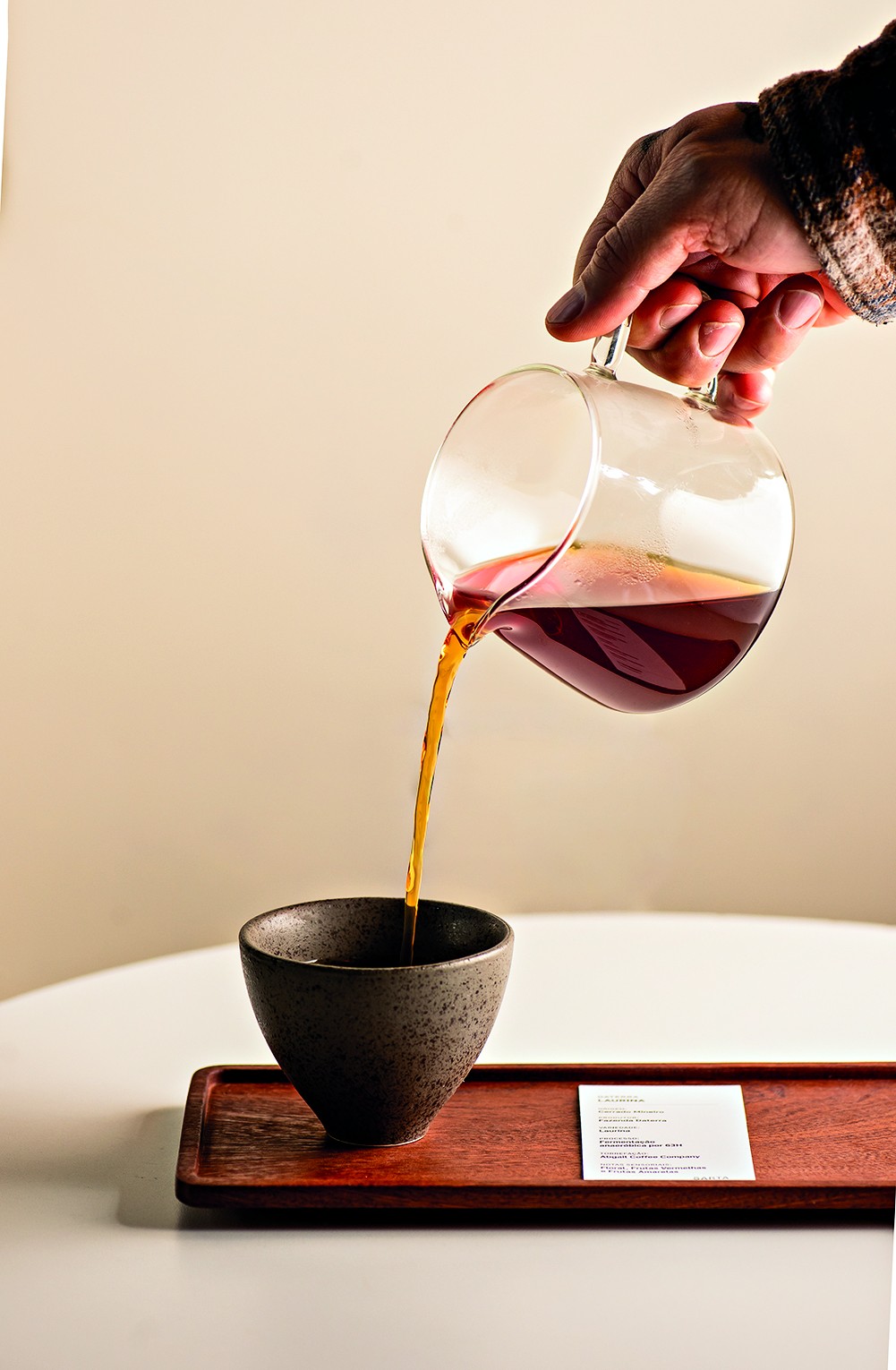 M&atilde;o de uma pessoa com manga xadrez derramando caf&eacute; coado de uma jarra de vidro transparente em uma x&iacute;cara de cer&acirc;mica escura, sobre uma bandeja de madeira com um cart&atilde;o branco de informa&ccedil;&otilde;es, em uma mesa branca