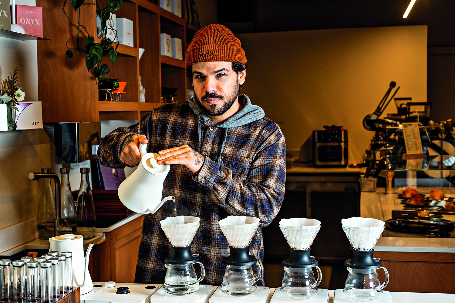 Homem barbudo com gorro laranja e camisa xadrez azul e marrom, concentrado, despejando &aacute;gua quente de uma chaleira branca em um coador de caf&eacute; sobre uma jarra de vidro. Outros coadores e jarras est&atilde;o alinhados em uma bancada clara, com prateleiras de madeira e equipamentos de caf&eacute; ao fundo