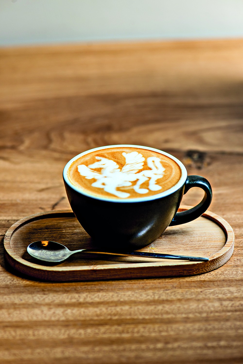 X&iacute;cara preta de caf&eacute; com arte latte de um unic&oacute;rnio branco, sobre um pires de madeira e uma colher, em uma mesa de madeira
