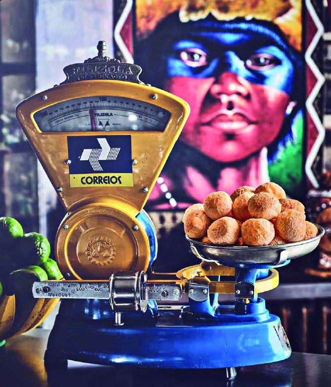 Balan&ccedil;a antiga Filizola amarela e azul com logo dos Correios, pesando p&atilde;es de queijo em um prato, com lim&otilde;es verdes &agrave; esquerda e um quadro de um ind&iacute;gena com pintura facial azul e vermelha ao fundo