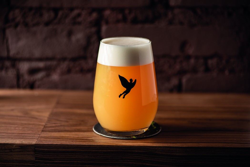 Copo de cerveja artesanal com l&iacute;quido &acirc;mbar e espuma branca, exibindo um adesivo preto de uma figura alada, sobre um descanso de copo met&aacute;lico em uma mesa de madeira escura, com parede de tijolos ao fundo