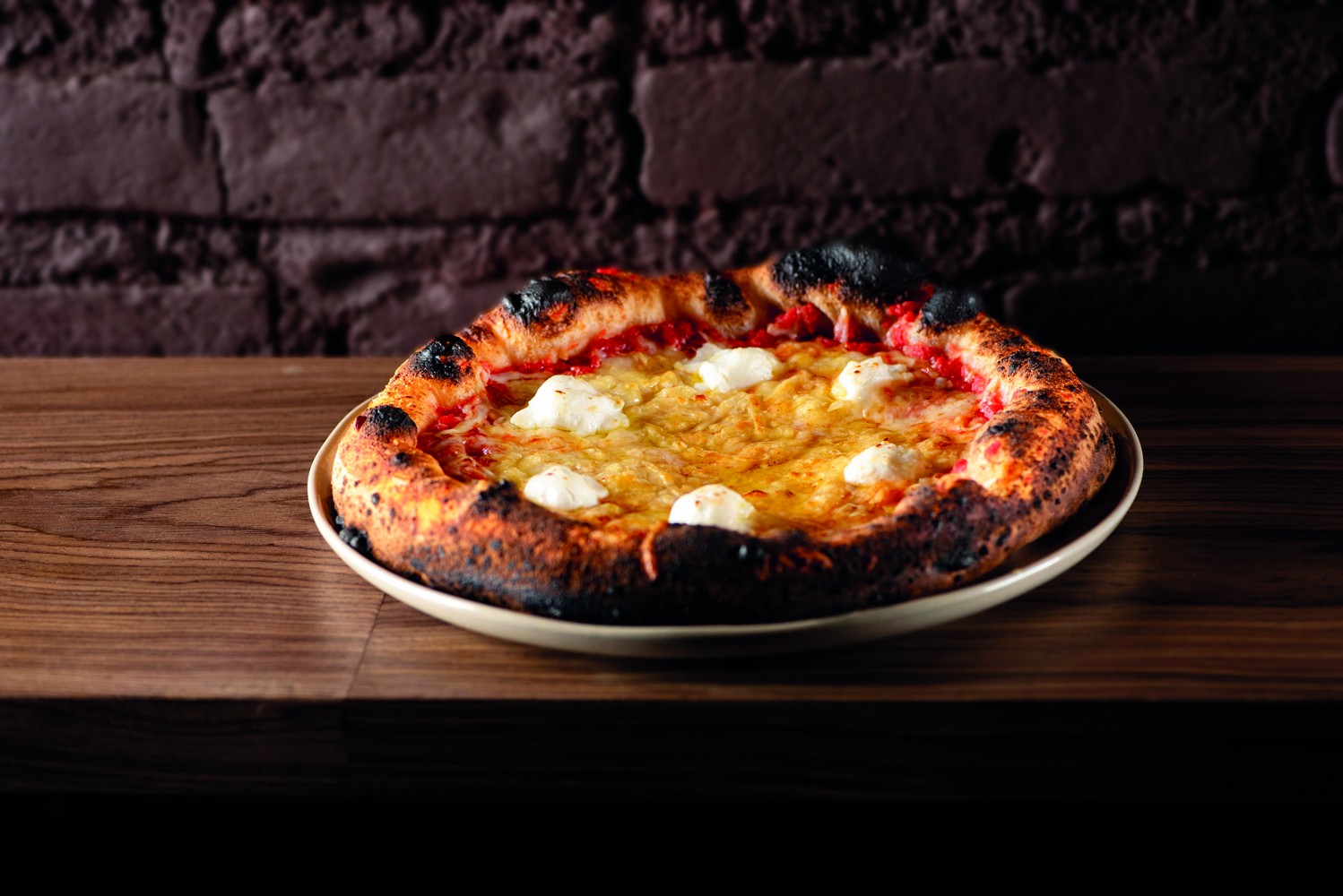 Pizza napolitana com borda alta e crocante, molho de tomate, queijo derretido e bolinhas de queijo branco, sobre um prato claro em uma mesa de madeira escura, com parede de tijolos ao fundo