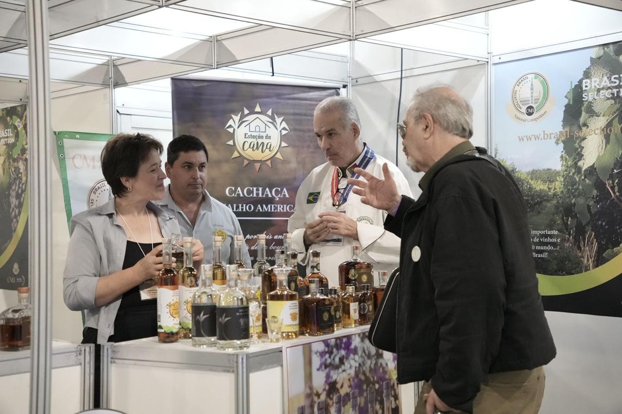 Quatro pessoas em um estande de exposi&ccedil;&atilde;o. Uma mulher de blusa cinza e um homem de camisa azul clara observam garrafas de cacha&ccedil;a. Um homem de jaleco branco e medalhas no pesco&ccedil;o conversa com um homem de casaco preto, que gesticula com a m&atilde;o direita. Ao fundo, banners de Esta&ccedil;&atilde;o da Cana e Brasil Selection