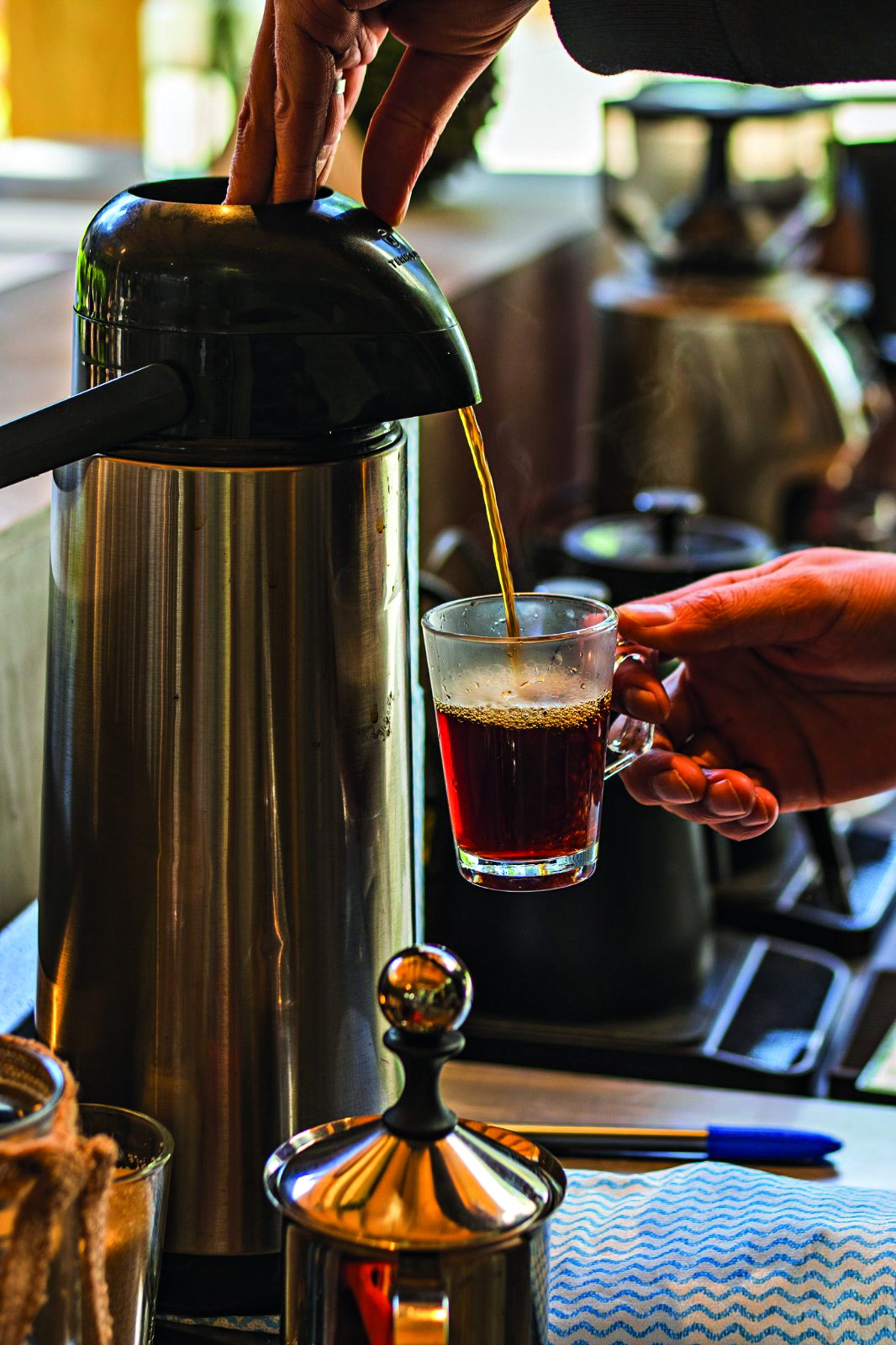 Uma pessoa serve caf&eacute; quente de uma garrafa t&eacute;rmica de inox em uma x&iacute;cara de vidro transparente, segurada pela outra m&atilde;o. O caf&eacute; forma um fio dourado, e a x&iacute;cara j&aacute; cont&eacute;m um pouco da bebida escura, com vapor subindo. Ao fundo, outros utens&iacute;lios de cozinha desfocados.