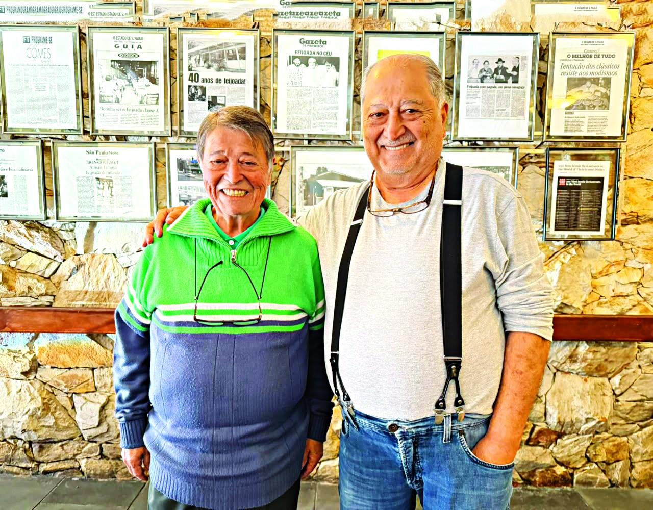 Dois homens idosos sorrindo para a c&acirc;mera, um de su&eacute;ter verde e azul com &oacute;culos pendurados, e outro de camiseta cinza, suspens&oacute;rios pretos e jeans, com &oacute;culos na gola, em frente a uma parede de pedra com quadros de jornais antigos