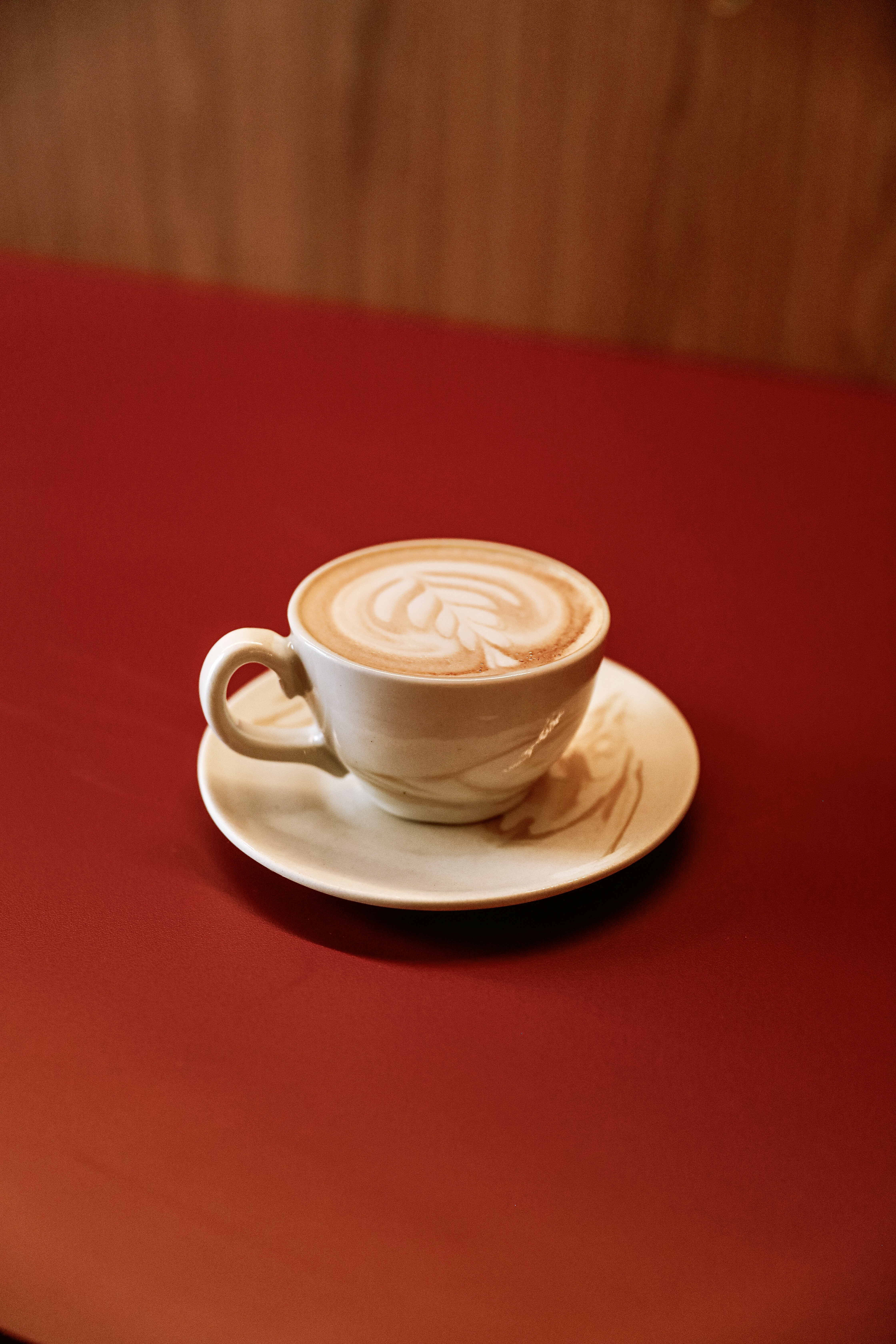 X&iacute;cara branca de caf&eacute; latte com arte de folha na espuma, sobre um pires branco, em uma mesa vermelha