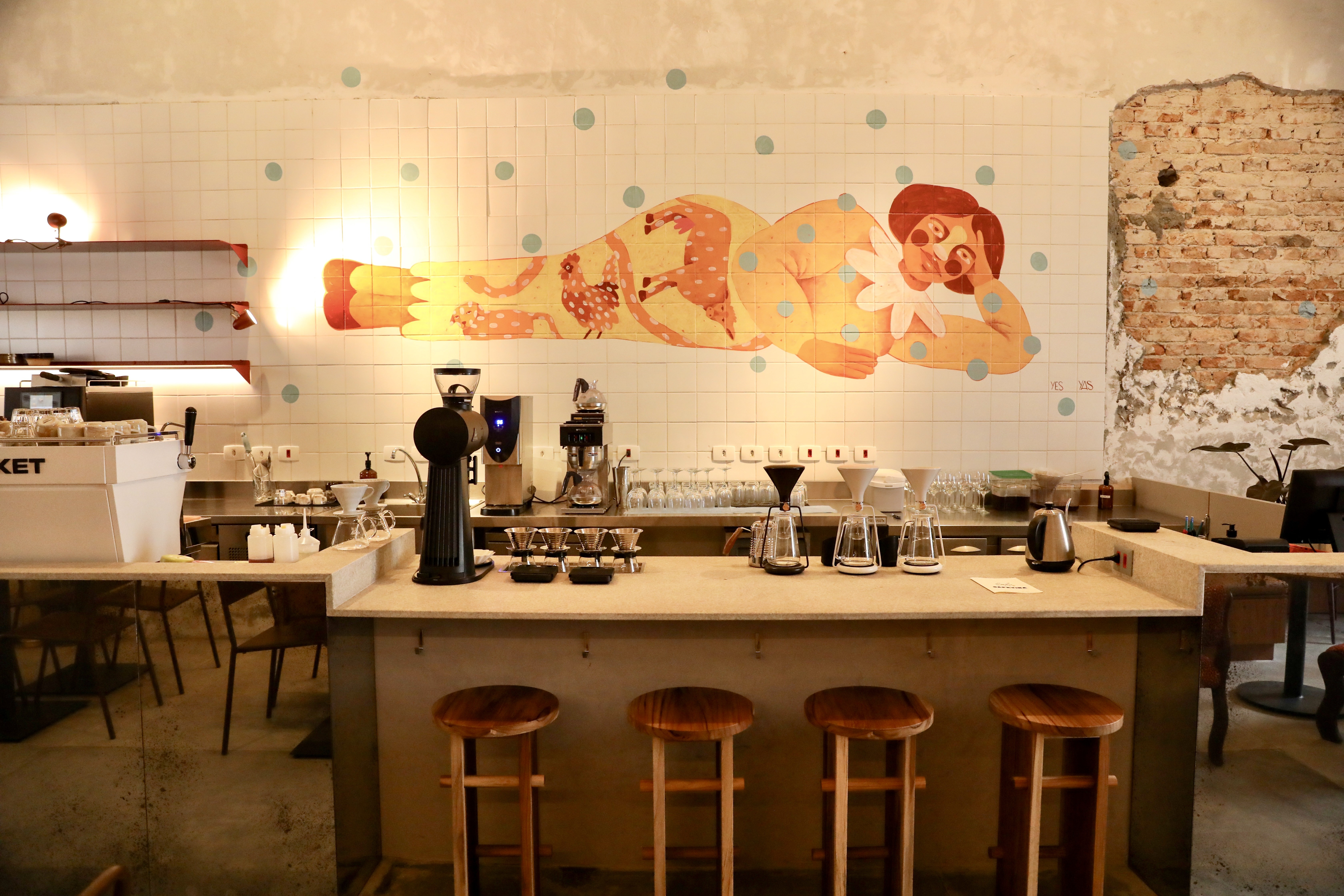 Interior de uma cafeteria com balc&atilde;o de madeira clara e quatro bancos altos de madeira. Ao fundo, uma parede de azulejos brancos com um mural de uma mulher deitada, em tons de amarelo e vermelho, e pontos azuis. Equipamentos de caf&eacute; e copos est&atilde;o sobre o balc&atilde;o. &Agrave; direita, uma parede de tijolos expostos e descascados.