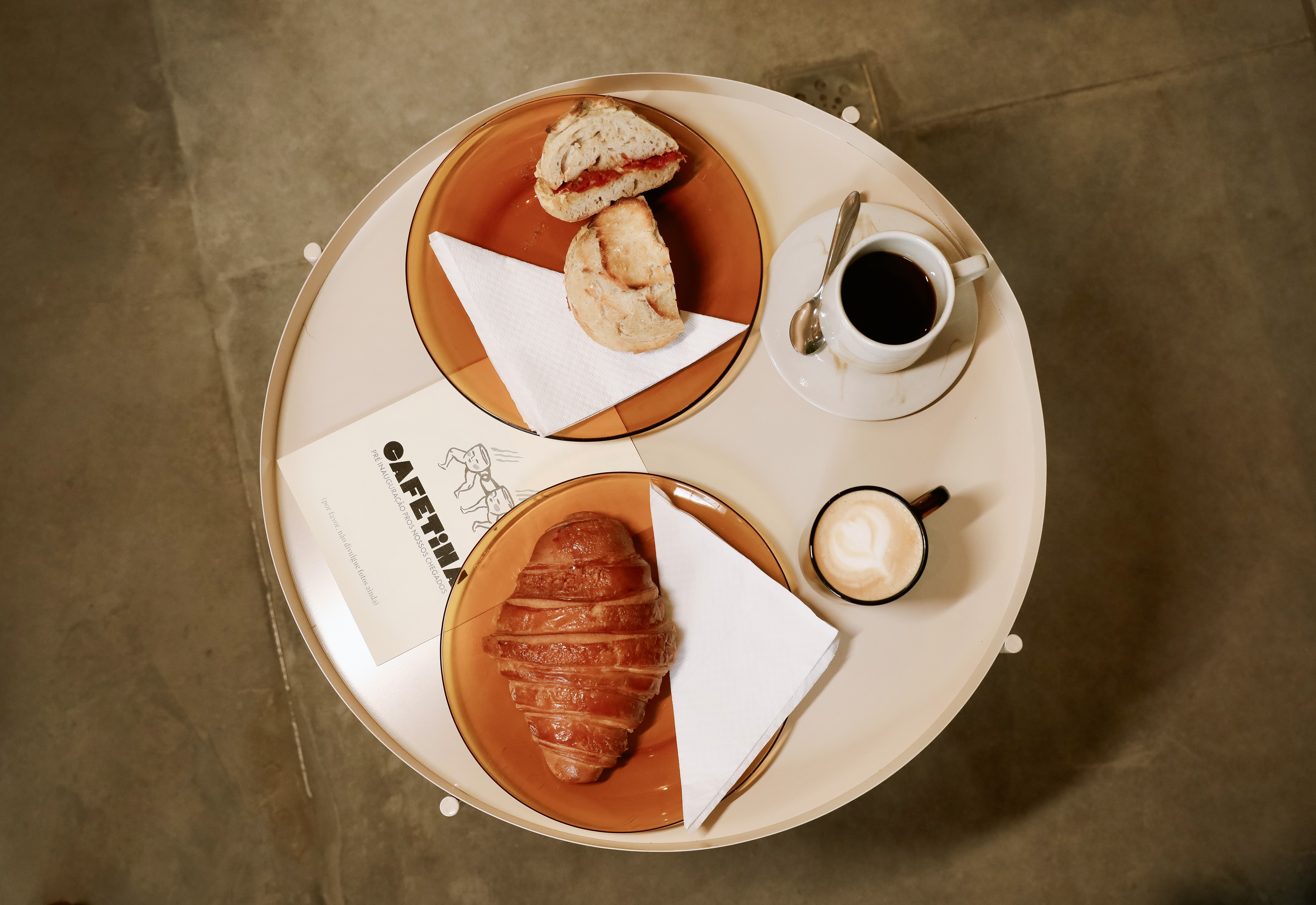 Vista a&eacute;rea de uma mesa redonda clara com caf&eacute; e lanches. H&aacute; um p&atilde;o com recheio vermelho e outro p&atilde;o simples em um prato, um croissant fatiado em outro, uma x&iacute;cara de caf&eacute; preto e uma de cappuccino com arte de cora&ccedil;&atilde;o, e um card&aacute;pio CAFETINA