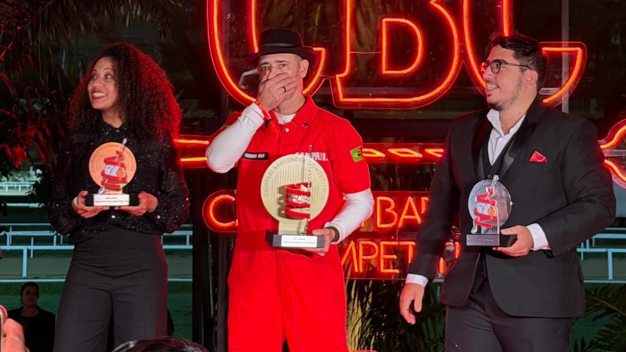 Tr&ecirc;s pessoas em um palco com letreiros de neon vermelhos ao fundo seguram trof&eacute;us. Uma mulher sorridente &agrave; esquerda, de blusa preta brilhante, segura um trof&eacute;u redondo. No centro, um homem de chap&eacute;u e macac&atilde;o vermelho cobre a boca com a m&atilde;o, segurando um trof&eacute;u dourado. &Agrave; direita, um homem de terno preto e &oacute;culos sorri, segurando um trof&eacute;u transparente.