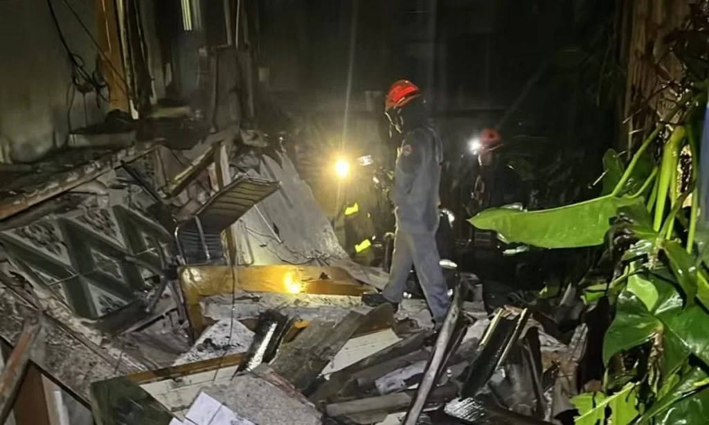 Equipe de resgate com capacetes e lanternas busca por sobreviventes em escombros de um prédio desabado, à noite, com vegetação ao lado