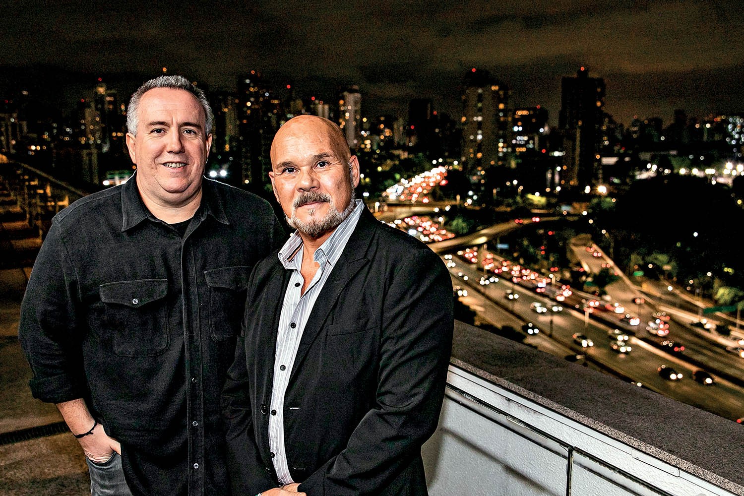 Dois homens sorrindo, um com camisa preta e outro de terno, posam em uma varanda com vista noturna para uma cidade iluminada e avenidas movimentadas