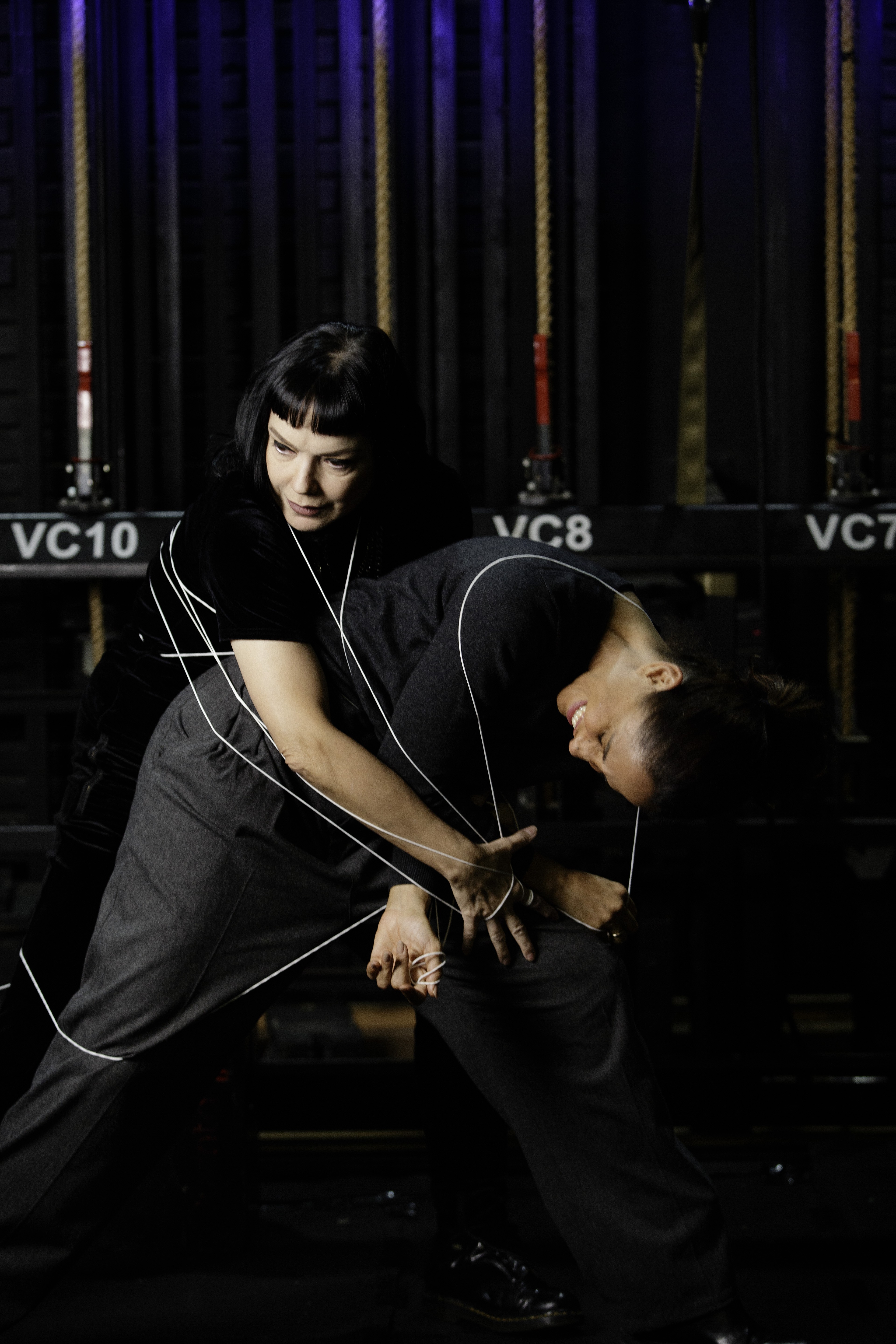 Duas mulheres em um palco escuro. Uma, de cabelo preto e franja, segura fios brancos que se estendem para a outra, que sorri com a cabe&ccedil;a inclinada para tr&aacute;s, como uma marionete. Ambas vestem roupas escuras e cal&ccedil;as. Ao fundo, estruturas de palco com as inscri&ccedil;&otilde;es VC10, VC8 e VC7