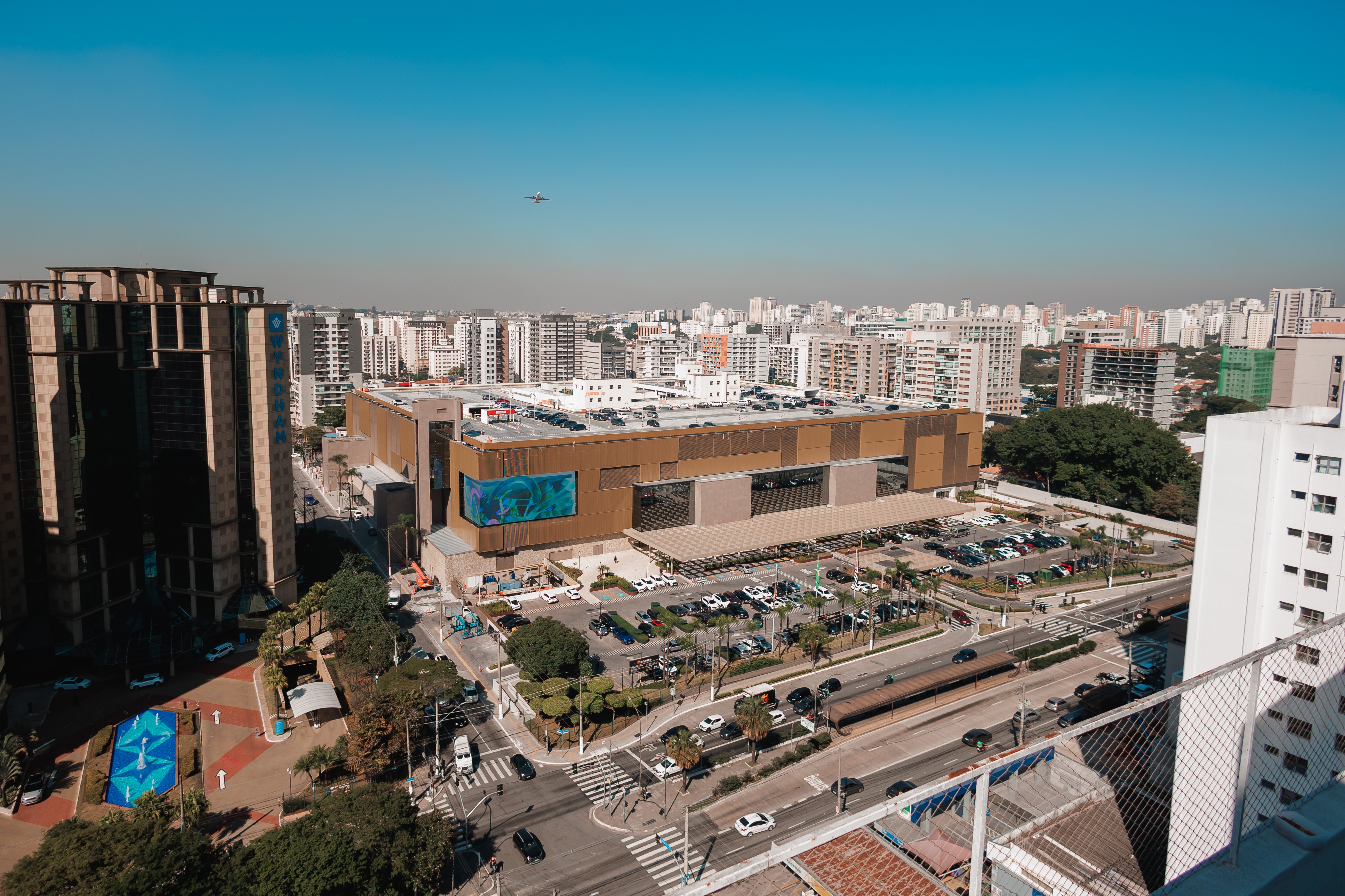 Vista aérea de uma cidade com um grande shopping center marrom no centro, rodeado por edifícios residenciais e comerciais. Há um estacionamento lotado, ruas com carros e um avião no céu azul claro. À esquerda, um prédio alto escuro e à direita, um edifício branco com uma grade de proteção no primeiro plano