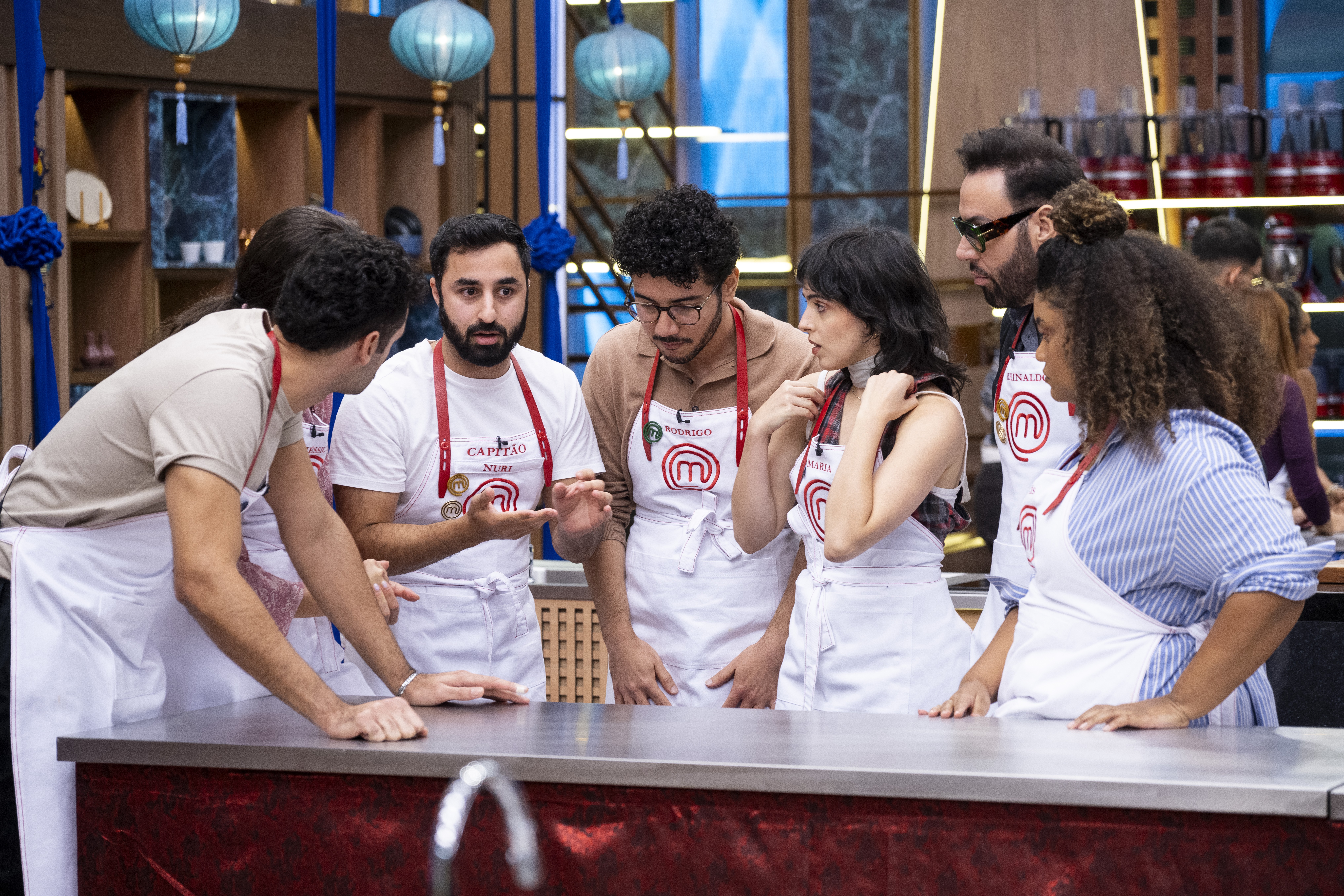 Grupo de sete pessoas, incluindo homens e mulheres, usando aventais brancos com detalhes vermelhos e o logo do MasterChef, reunidos em uma bancada de cozinha. Eles parecem estar em uma competi&ccedil;&atilde;o culin&aacute;ria, conversando e gesticulando, com um fundo de estantes e lumin&aacute;rias azuis