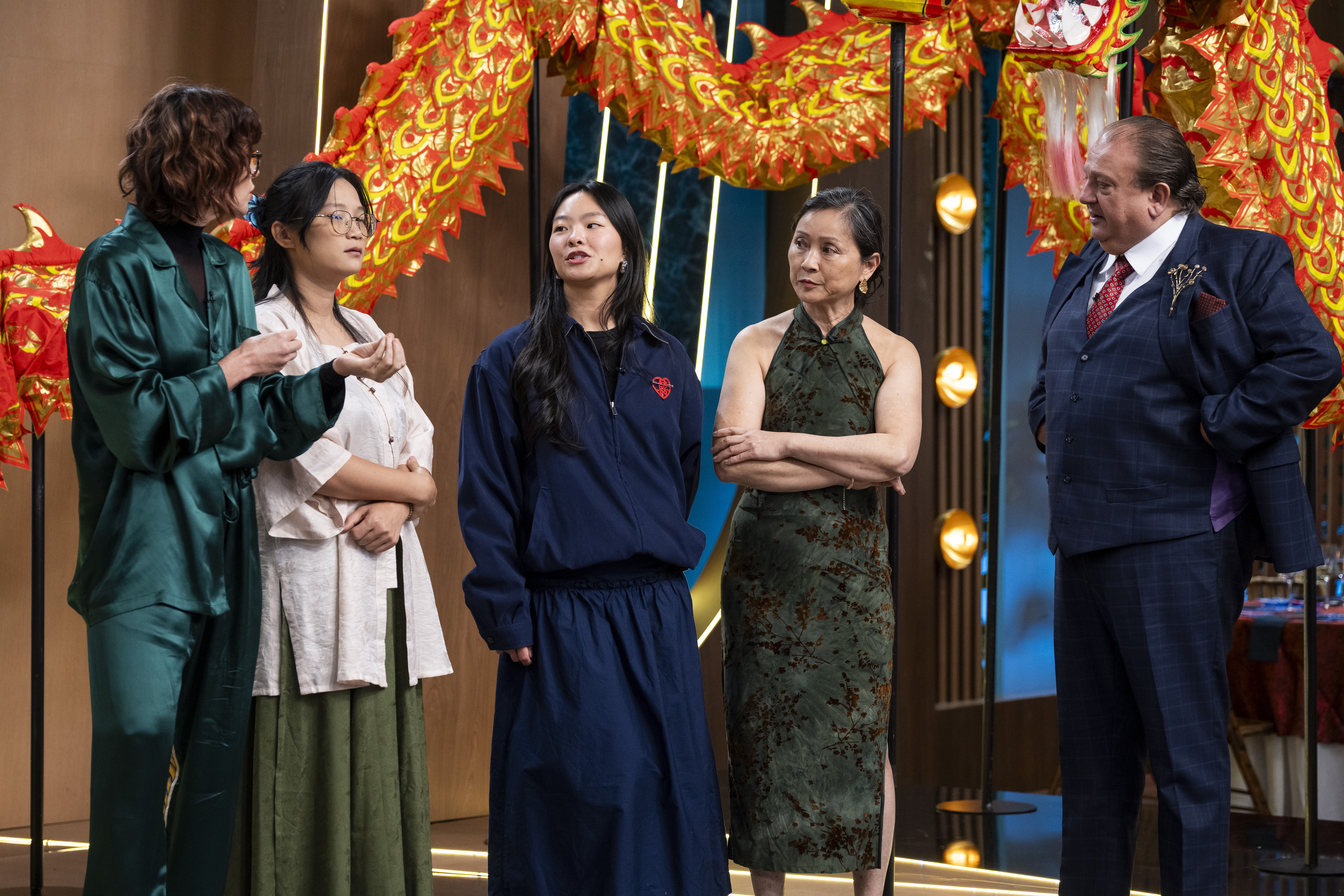 Cinco pessoas, tr&ecirc;s mulheres e dois homens, em um cen&aacute;rio com decora&ccedil;&otilde;es de drag&otilde;es chineses. &Agrave; esquerda, uma mulher de cabelo cacheado e camisa verde-esmeralda conversa com uma mulher de &oacute;culos, camisa branca e saia verde-oliva. Ao centro, uma mulher de cabelo liso e conjunto azul-marinho olha para cima. &Agrave; direita, uma mulher de vestido verde estampado e bra&ccedil;os cruzados observa um homem de terno azul-marinho e gravata vermelha, que parece falar com ela