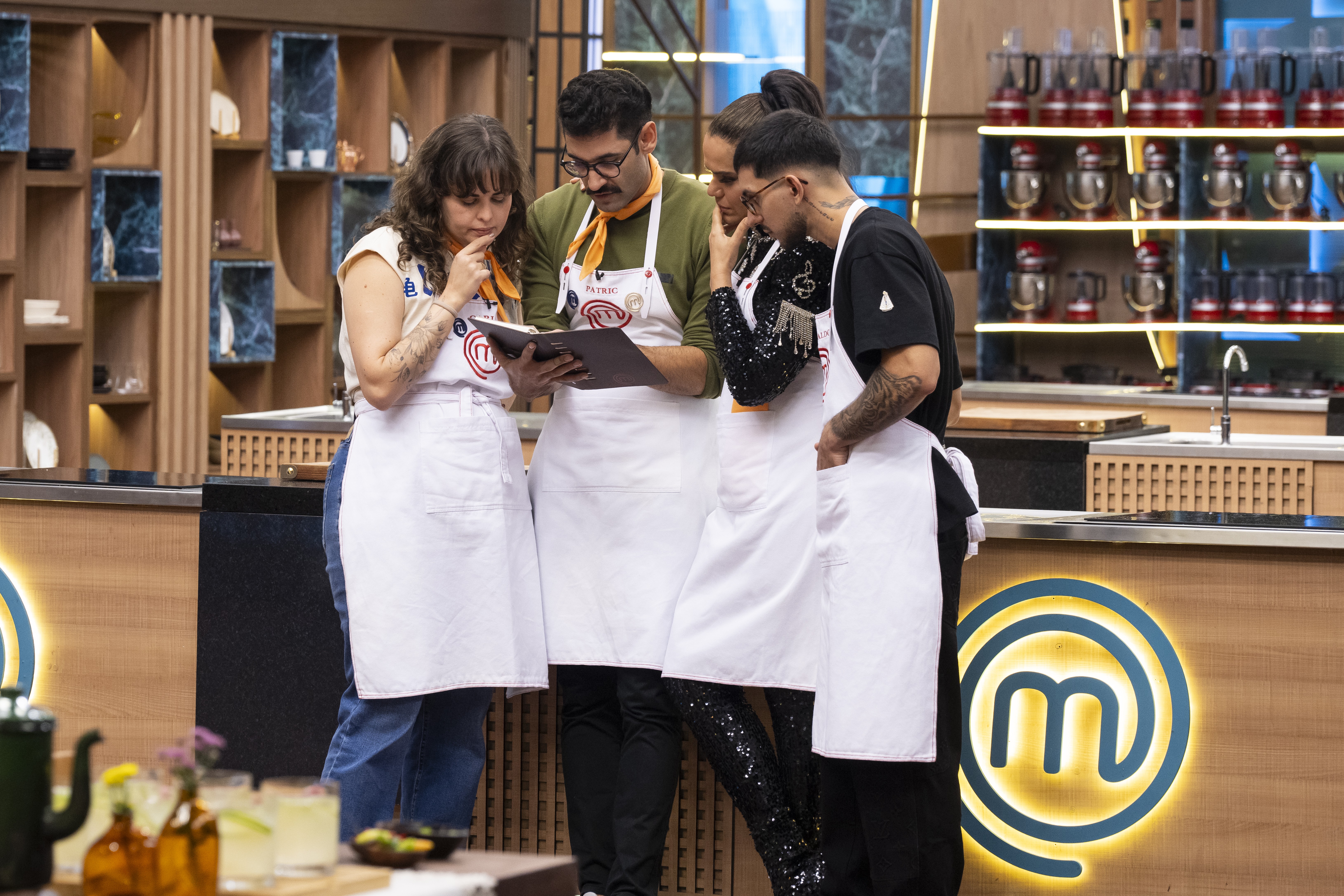 Quatro chefs, tr&ecirc;s homens e uma mulher, vestindo aventais brancos, olham atentamente para um caderno na cozinha do MasterChef. A mulher &agrave; esquerda tem a m&atilde;o no queixo, pensativa. O homem central, de bigode e &oacute;culos, segura o caderno. Ao fundo, prateleiras de madeira com objetos decorativos e utens&iacute;lios de cozinha. O logo do MasterChef brilha na bancada.
