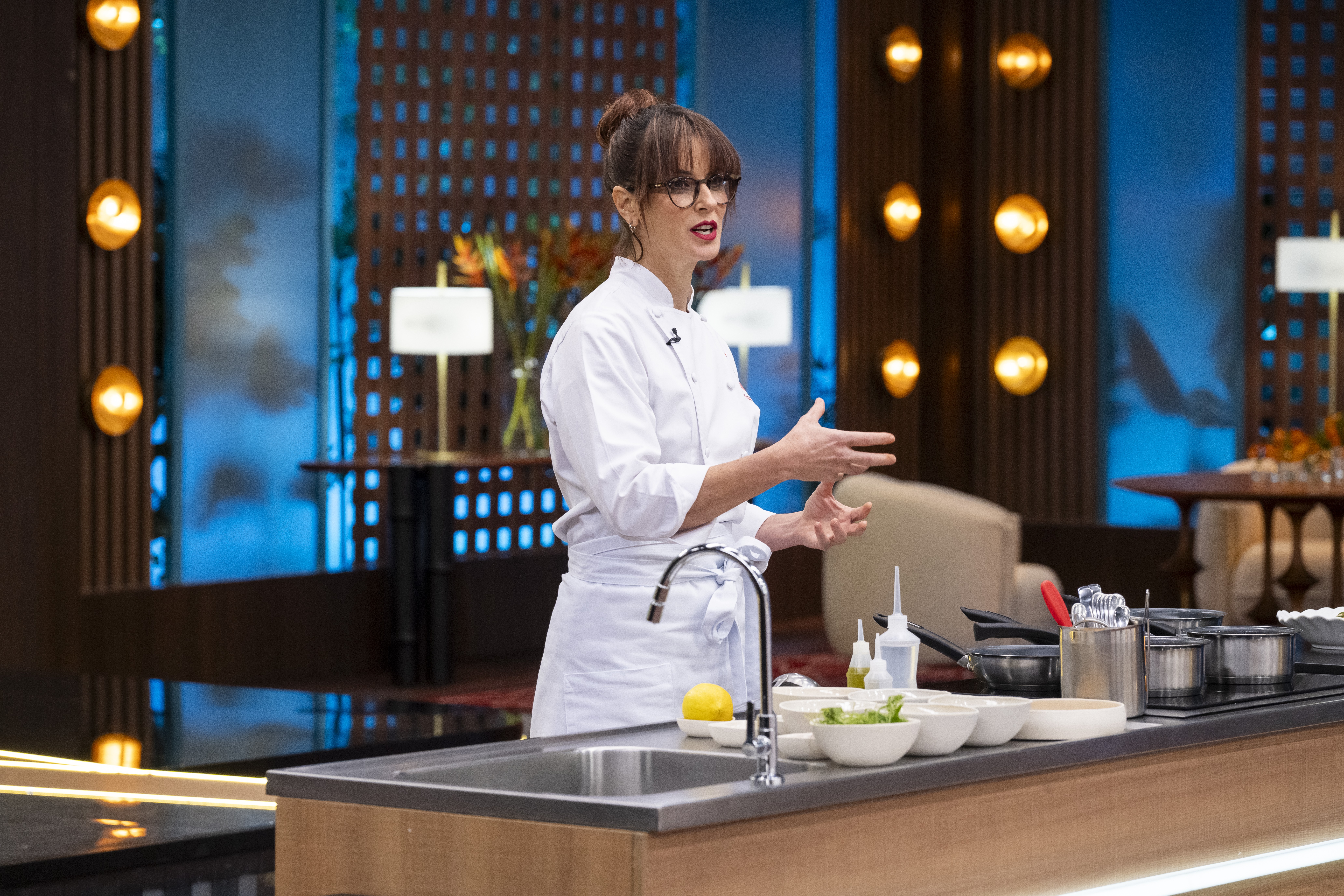 Mulher de &oacute;culos, cabelo preso e uniforme de chef branco, gesticula com as m&atilde;os abertas em um est&uacute;dio de cozinha. Ela est&aacute; atr&aacute;s de uma bancada com pia, lim&atilde;o, tigelas e panelas. Ao fundo, pain&eacute;is de madeira com luzes douradas e um ambiente com mesas e cadeiras.