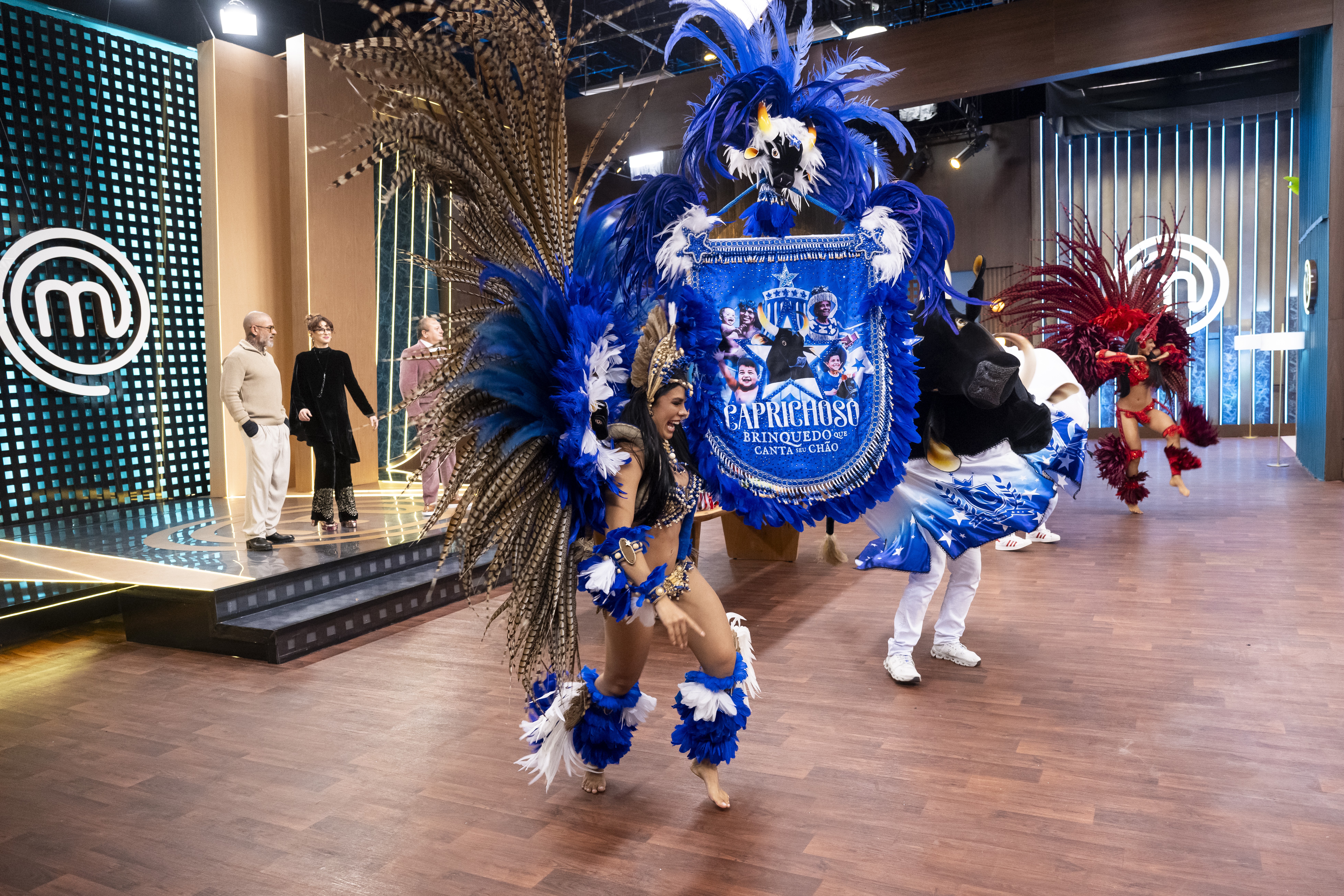 Mulher sorridente em traje de carnaval azul e marrom com plumas, dan&ccedil;ando no centro de um palco. Ao fundo, tr&ecirc;s pessoas assistem, e &agrave; direita, outra dan&ccedil;arina de vermelho.