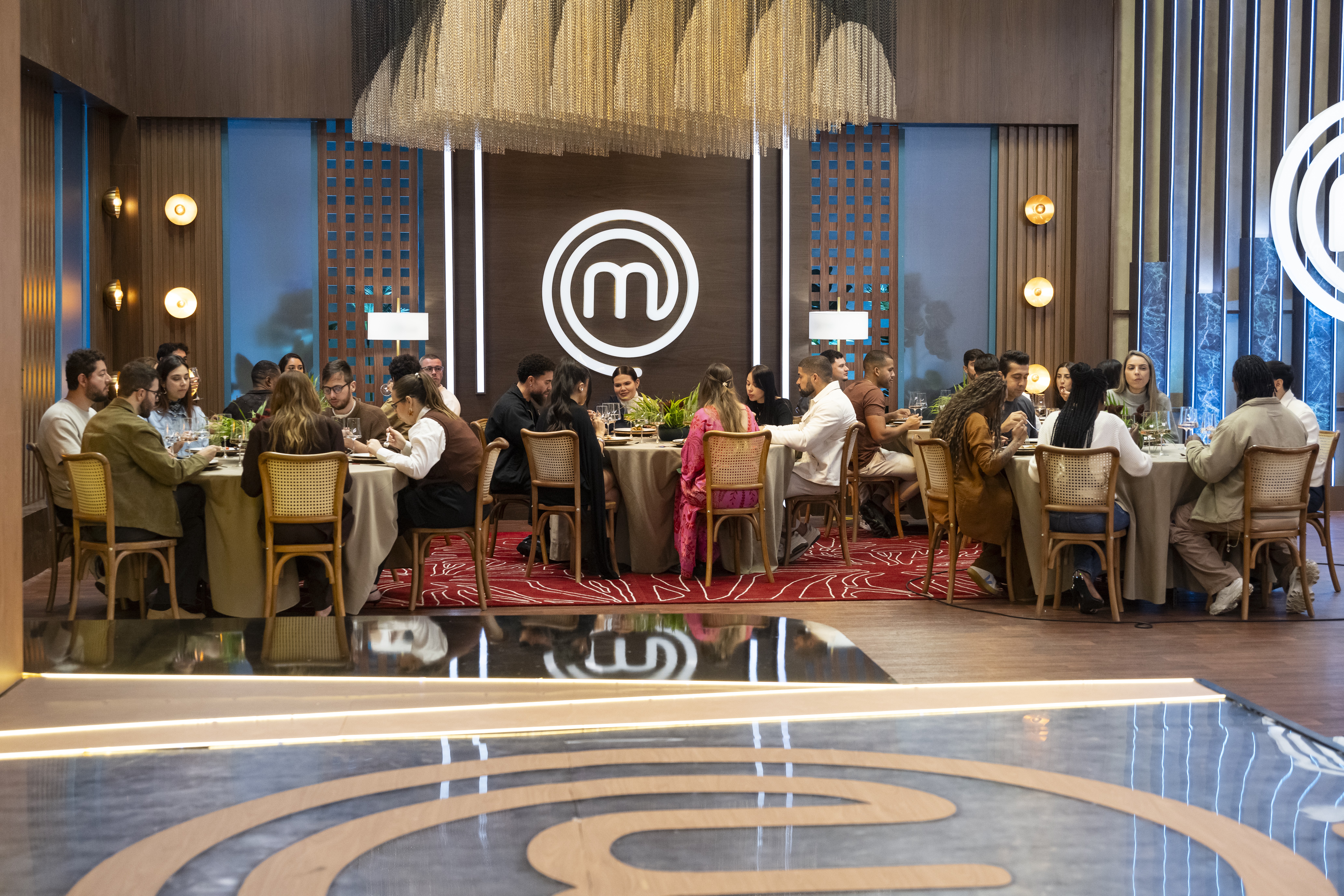 Pessoas sentadas em mesas redondas, comendo e conversando, em um ambiente de restaurante com ilumina&ccedil;&atilde;o aconchegante e o logo do MasterChef ao fundo