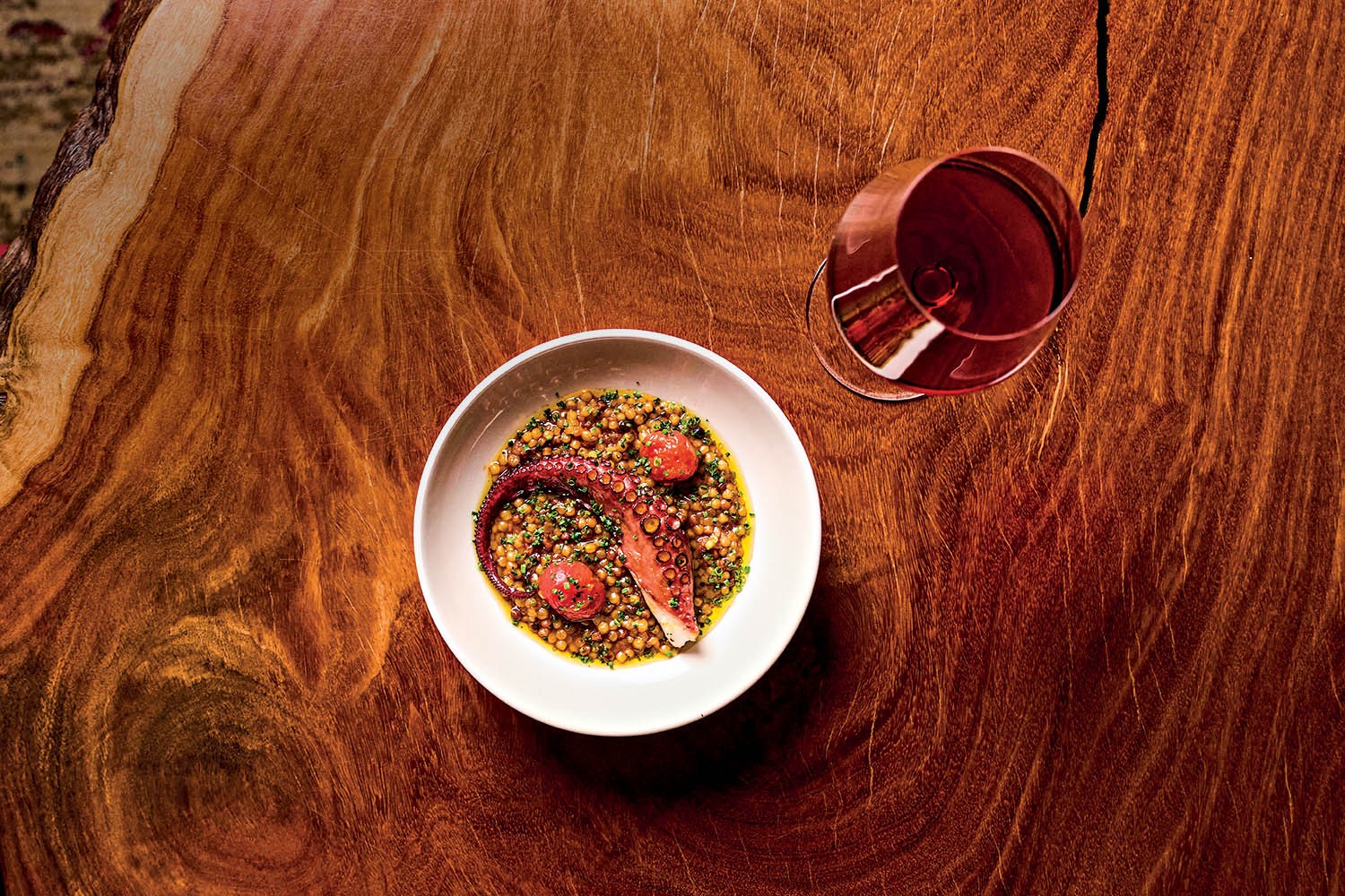 Prato branco com tent&aacute;culo de polvo, cuscuz marroquino, ervas e tomates-cereja, ao lado de uma ta&ccedil;a de vinho tinto, sobre uma mesa de madeira r&uacute;stica com veios marcantes