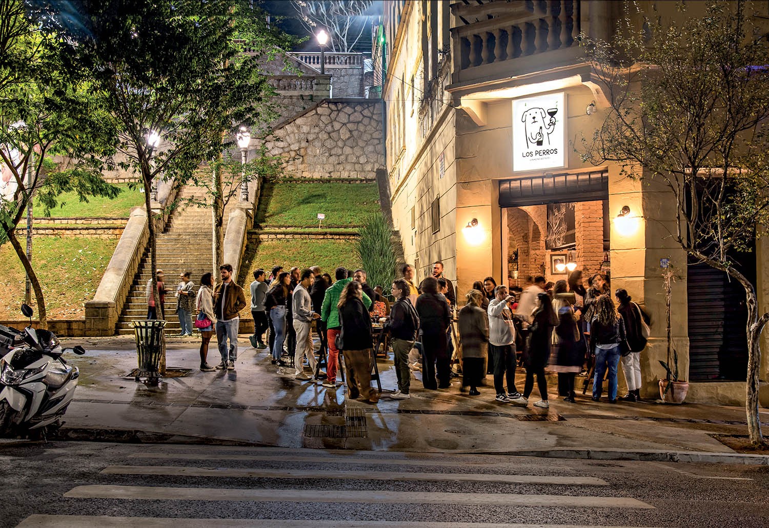 Grupo de pessoas conversando e bebendo em frente a um bar iluminado &agrave; noite, com uma escadaria de pedra e &aacute;rvores ao fundo. Uma motocicleta branca est&aacute; estacionada &agrave; esquerda e uma faixa de pedestres cruza a rua em primeiro plano