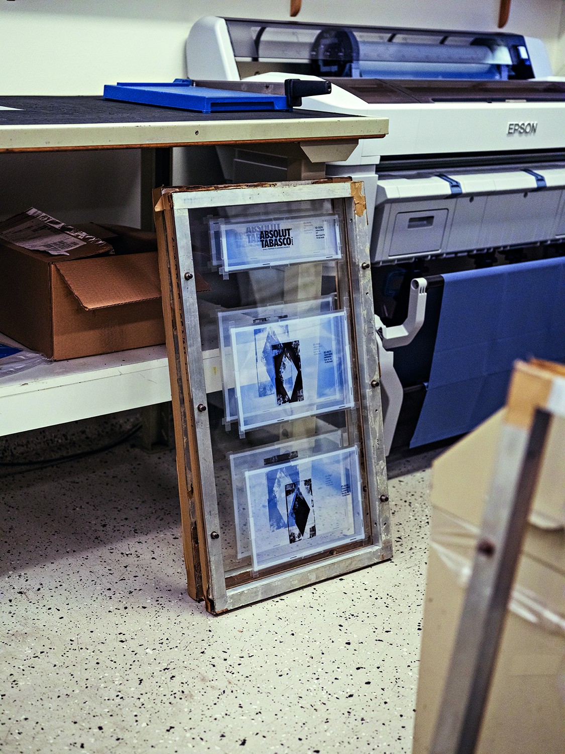 Impressora Epson de grande formato, com um expositor de acr&iacute;lico e madeira contendo impress&otilde;es de Absolut Tabasco e outras artes gr&aacute;ficas, em um ambiente de trabalho com piso claro e salpicado