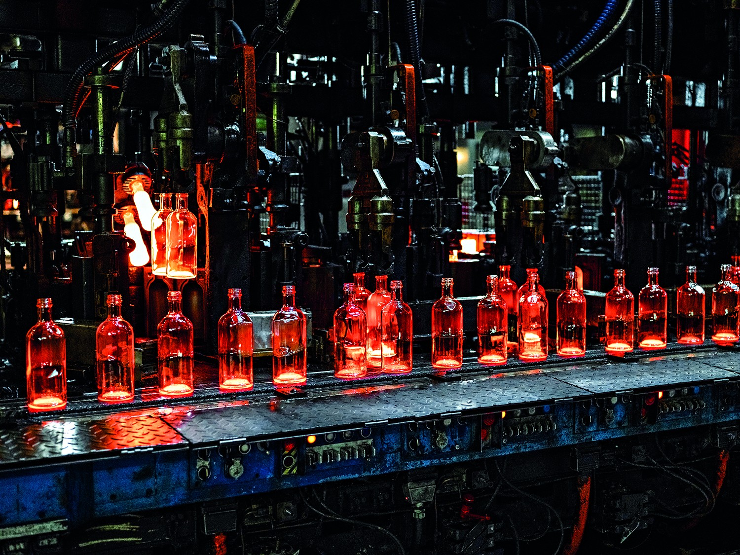 Garrafas de vidro incandescentes, em tons de laranja e vermelho brilhante, sendo transportadas por uma esteira em uma f&aacute;brica escura. M&aacute;quinas e tubula&ccedil;&otilde;es complexas dominam o fundo, com algumas garrafas ainda em processo de moldagem, emitindo um brilho intenso.