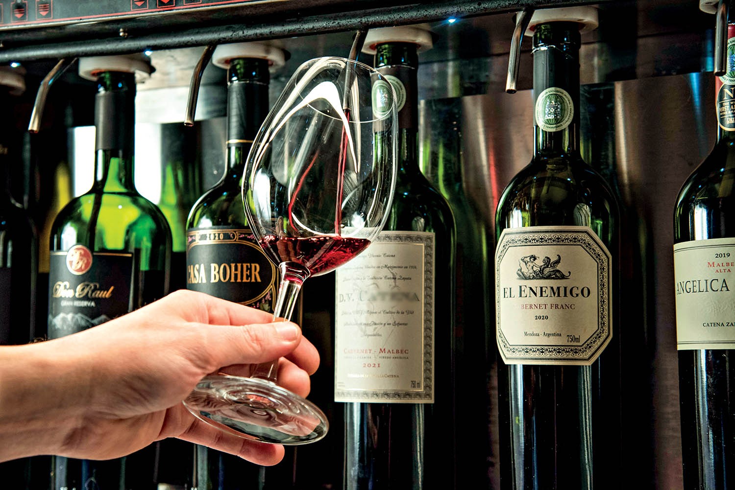 Uma m&atilde;o segura uma ta&ccedil;a de vinho tinto, com v&aacute;rias garrafas de vinho em um dispensador ao fundo. As garrafas t&ecirc;m r&oacute;tulos como El Enemigo e Angelica