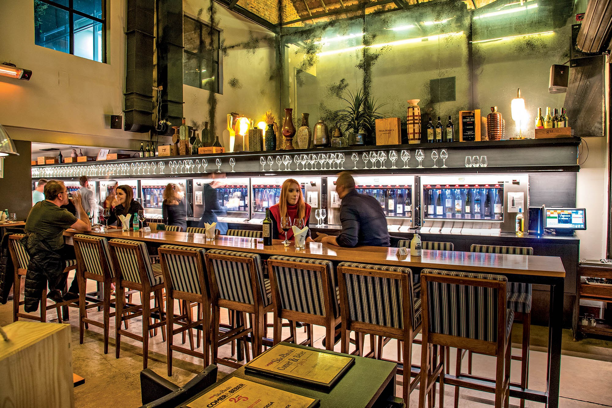 Pessoas sentadas em bancos altos em um bar de vinhos com garrafas expostas em prateleiras iluminadas ao fundo, criando um ambiente aconchegante