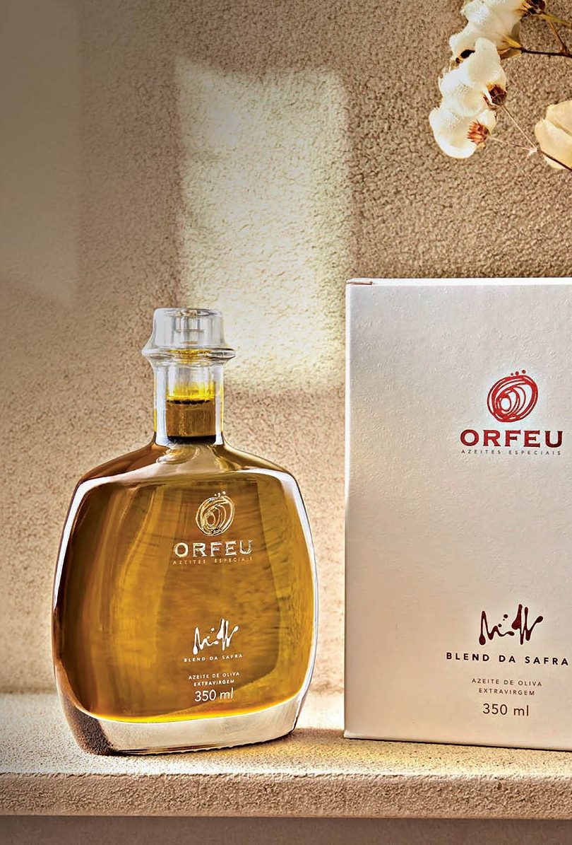 Garrafa de azeite Orfeu de 350 ml, com l&iacute;quido dourado, ao lado de sua caixa branca com o mesmo logo, sobre uma superf&iacute;cie clara. Ao fundo, uma parede texturizada e galhos secos com flores brancas, iluminados por um feixe de luz
