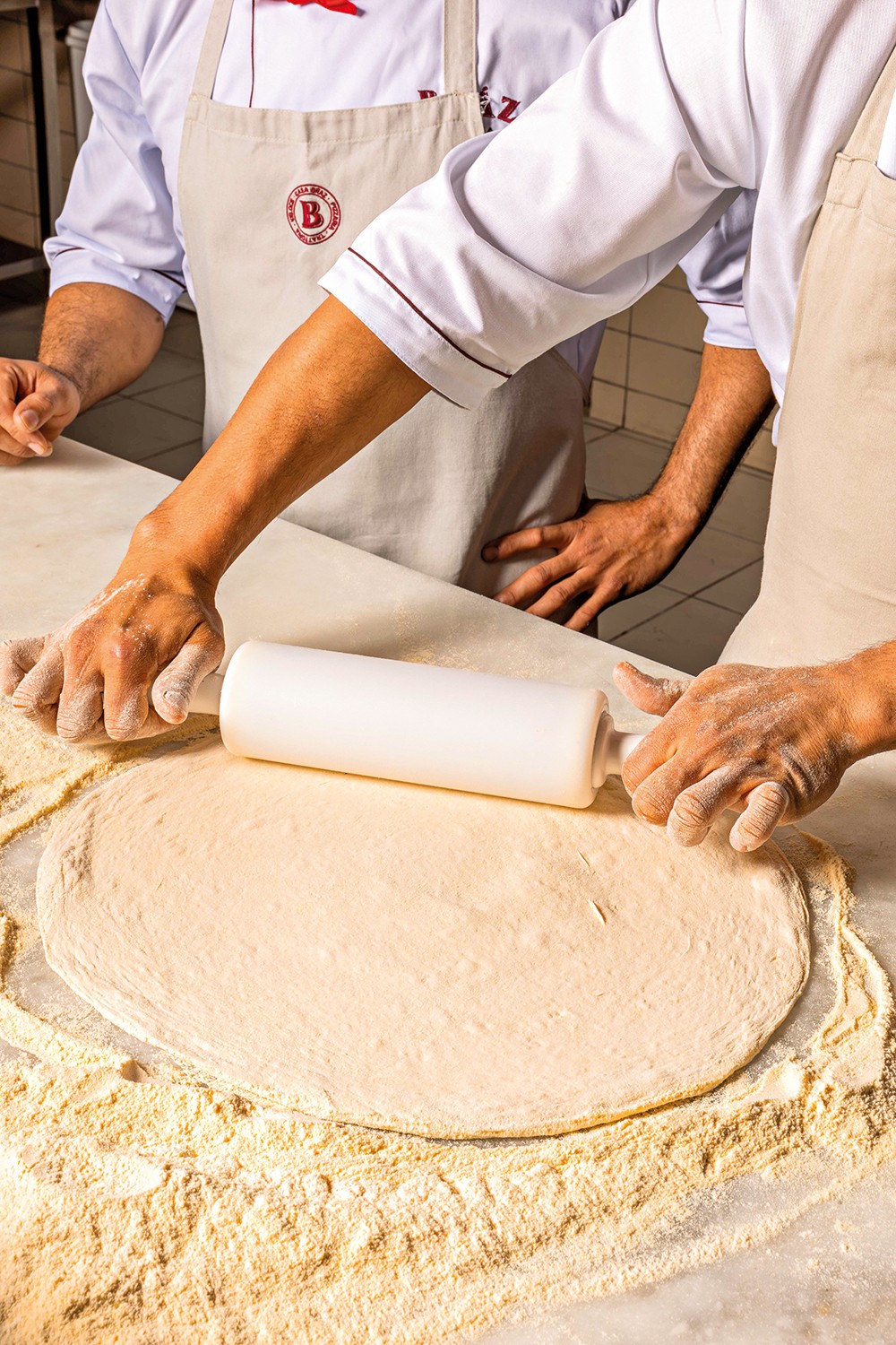 Duas pessoas, vestindo aventais e uniformes de chef, esticam uma massa de pizza com um rolo de massa branco sobre uma bancada enfarinhada