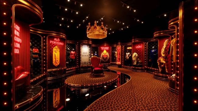 Exposi&ccedil;&atilde;o luxuosa com paredes vermelhas e douradas, tapete estampado de on&ccedil;a, uma coroa dourada gigante no teto e uma poltrona vermelha no centro. Pain&eacute;is exibem manequins, objetos e letreiros luminosos como AL&Ocirc; PORTEIRO e LE&Atilde;O